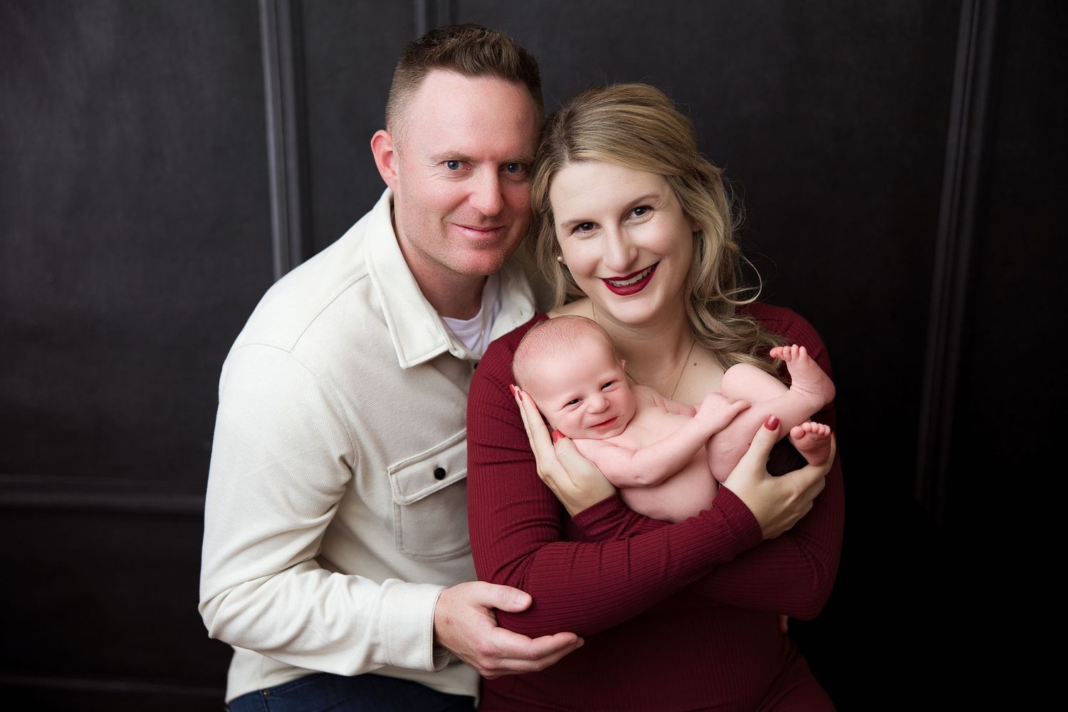 A family poses together in a studio portrait against a dark background wearing white and burgundy clothing.