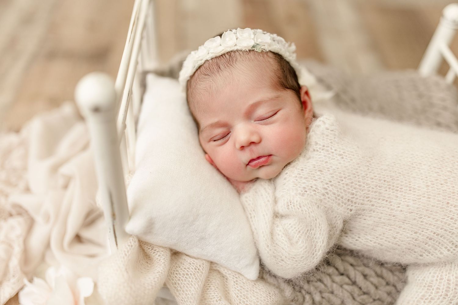 Sweet sleeping newborn wearing lace headband and white knit outfit on delicate bedding.