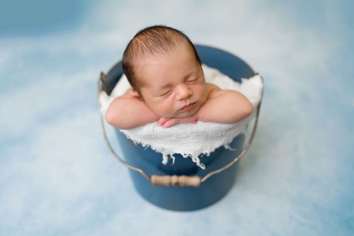 Newborn peacefully sleeps in round blue bowl against soft blue backdrop.