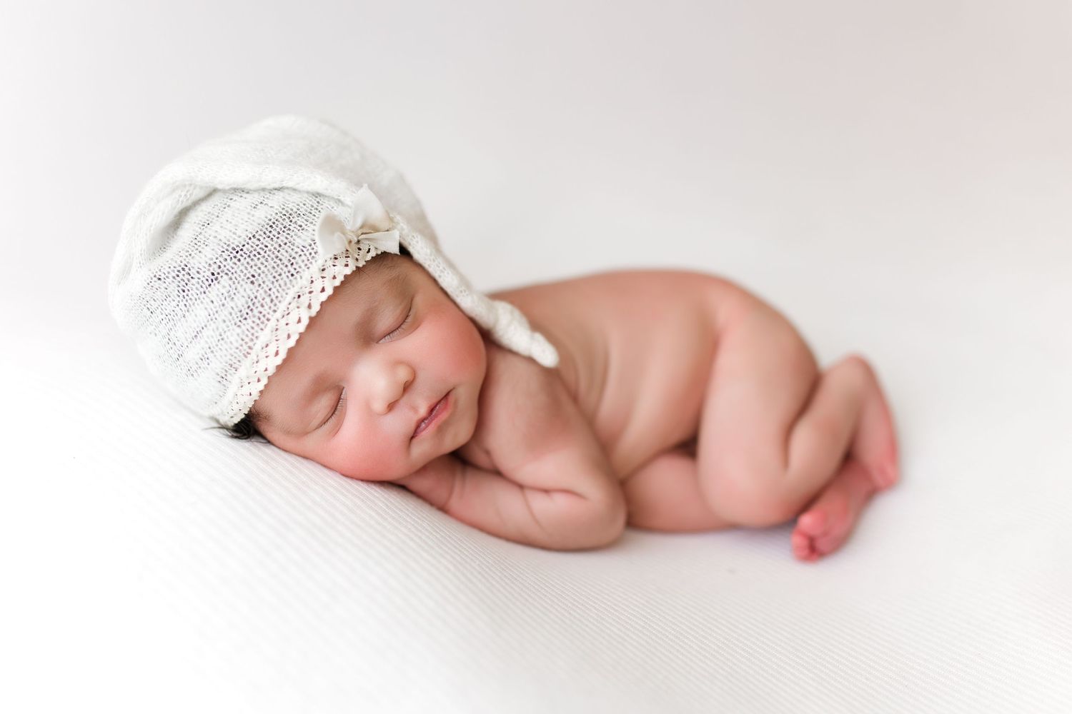 Infant wearing white resting on soft white blanket in minimalist newborn photography setup.