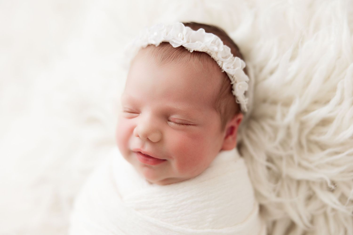 Peaceful sleeping newborn baby wrapped in white with lace headband on fluffy white blanket.