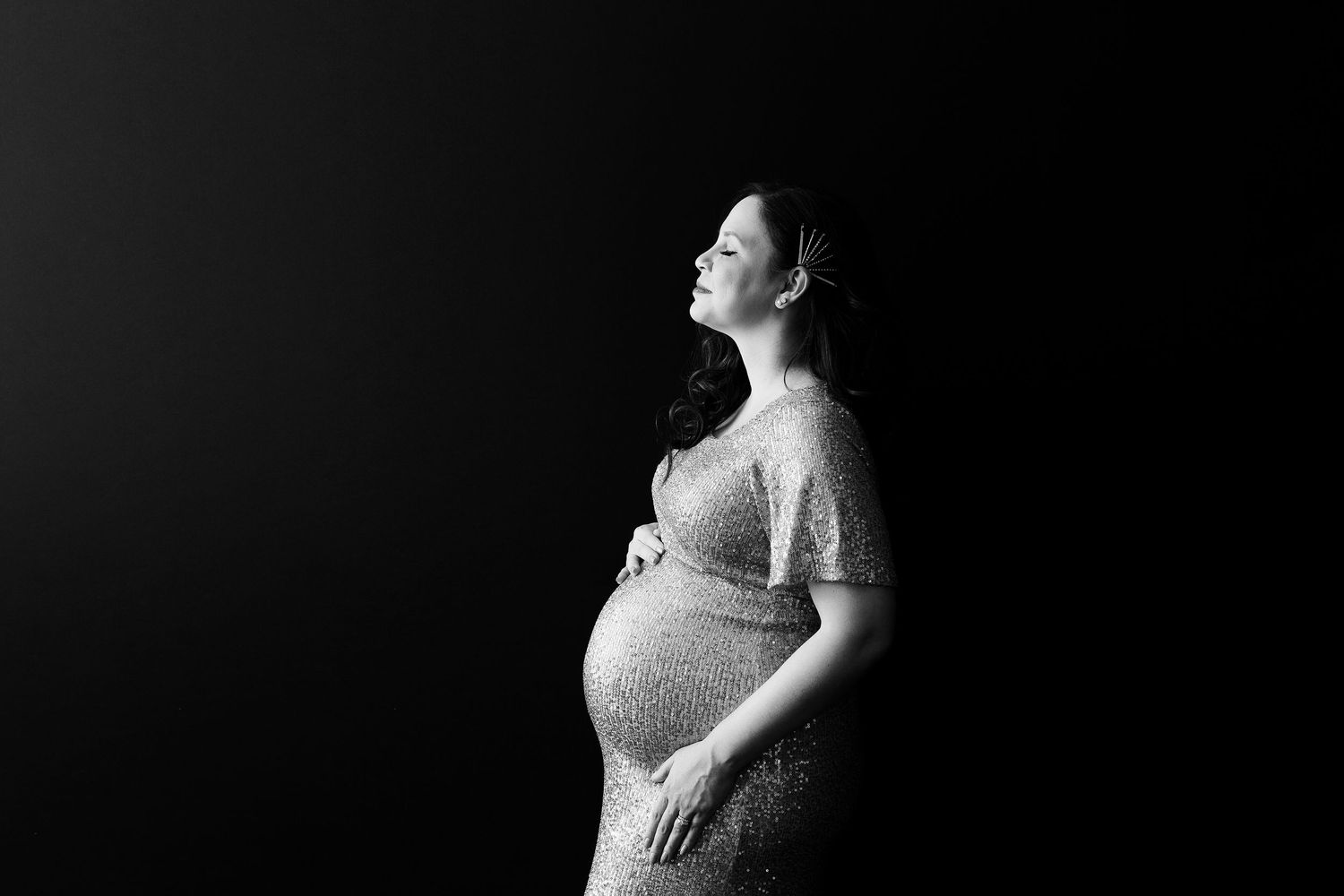 Dramatic side profile maternity portrait in sequined dress against black background.