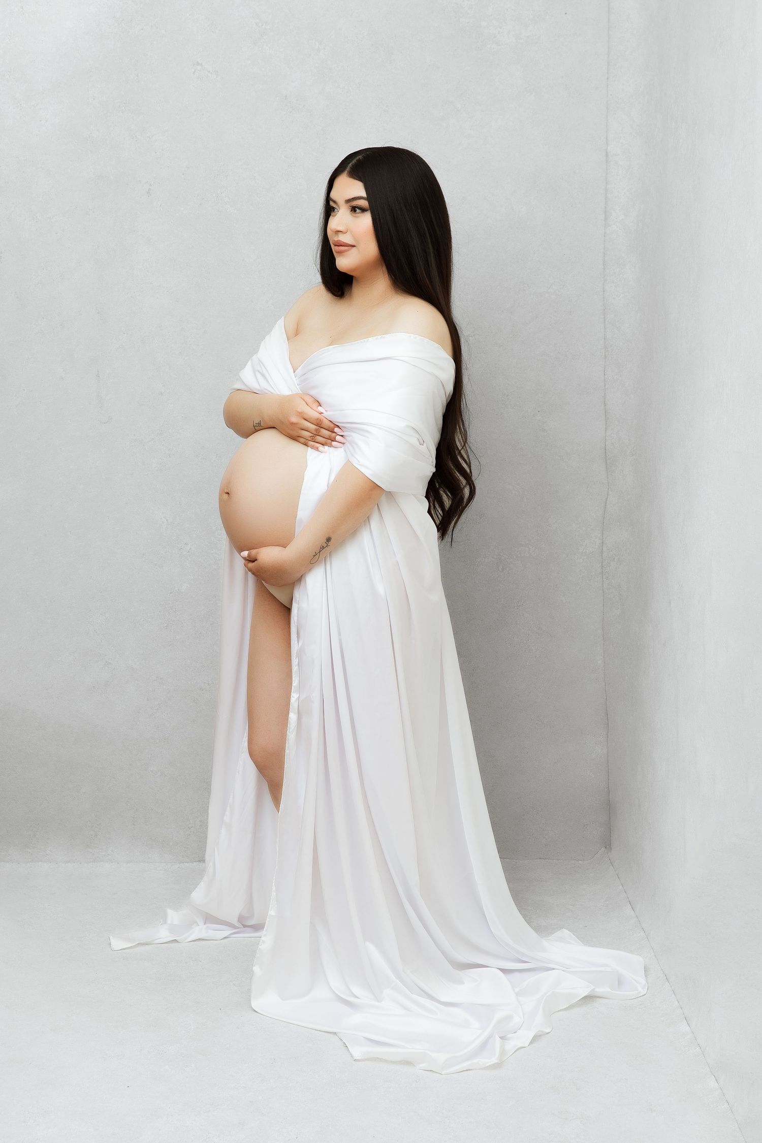 Maternity portrait in elegant white flowing dress against grey backdrop.