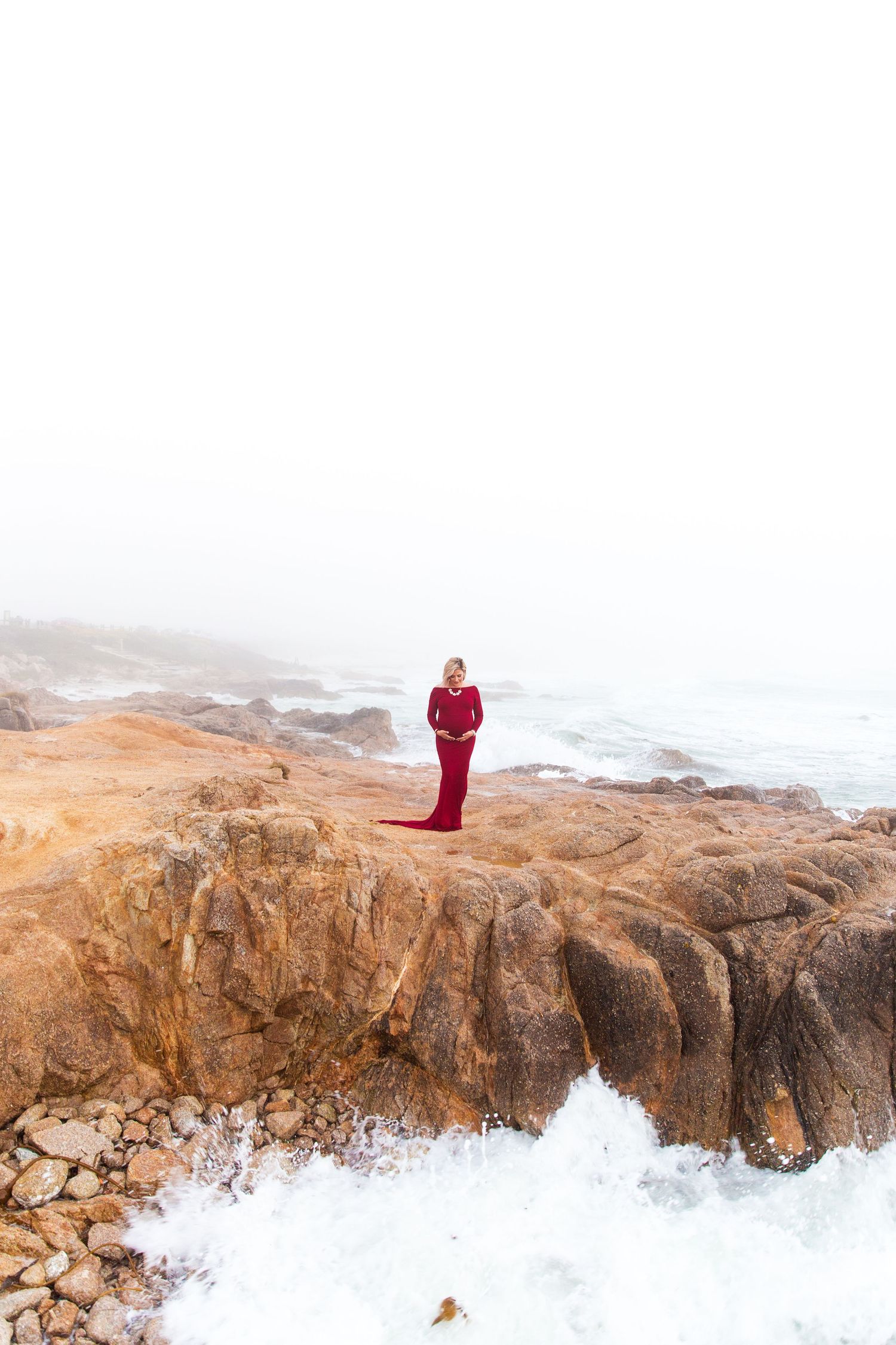 Person in red dress standing on rocky coastline with crashing waves below.