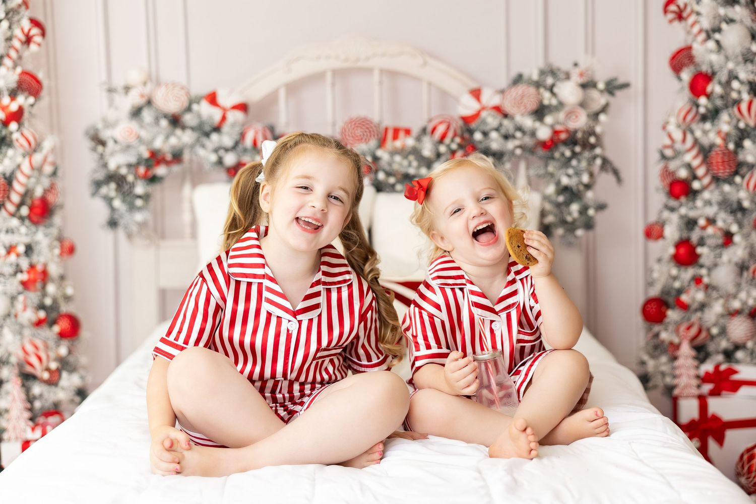Two children in matching striped pajamas sit on white bedding with red and white Christmas background, milk and cookie mini sessions.