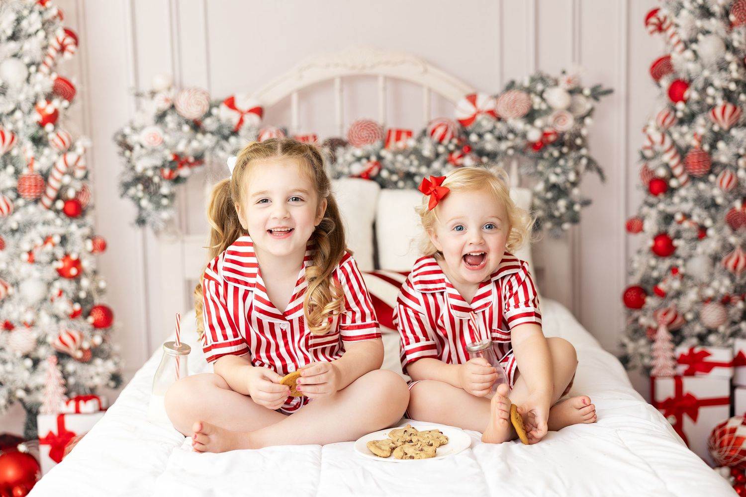 Two children in matching red striped pajamas sitting on white bed with Christmas decorations.