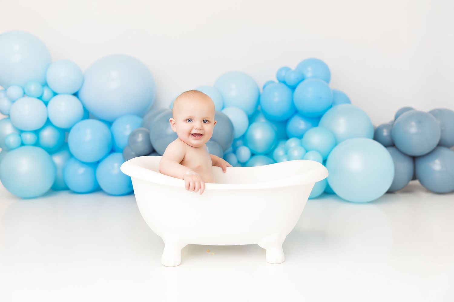 Baby in white bathtub with blue balloon arch backdrop during professional photo session.