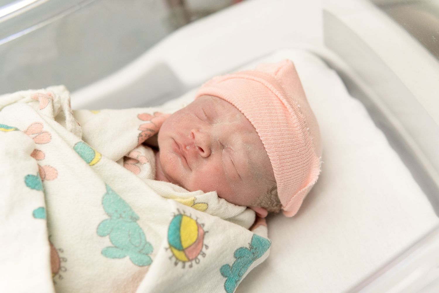 A sleeping baby in pink lies on white bedding with colorful bunny-themed nursery decor visible.