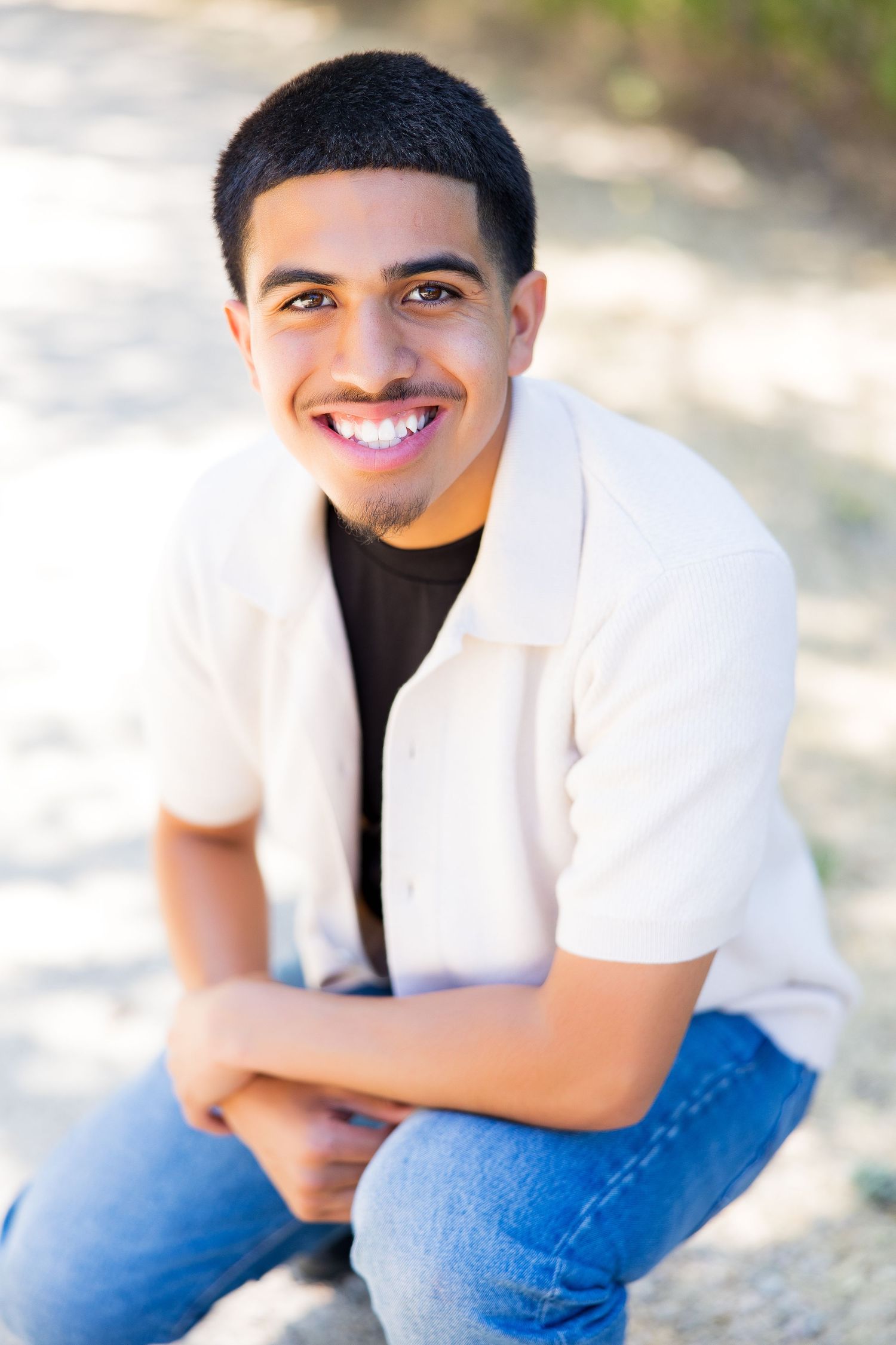 Young Hispanic man smiling outdoors, wearing white shirt over black tee with jeans, sitting relaxed in natural setting.
