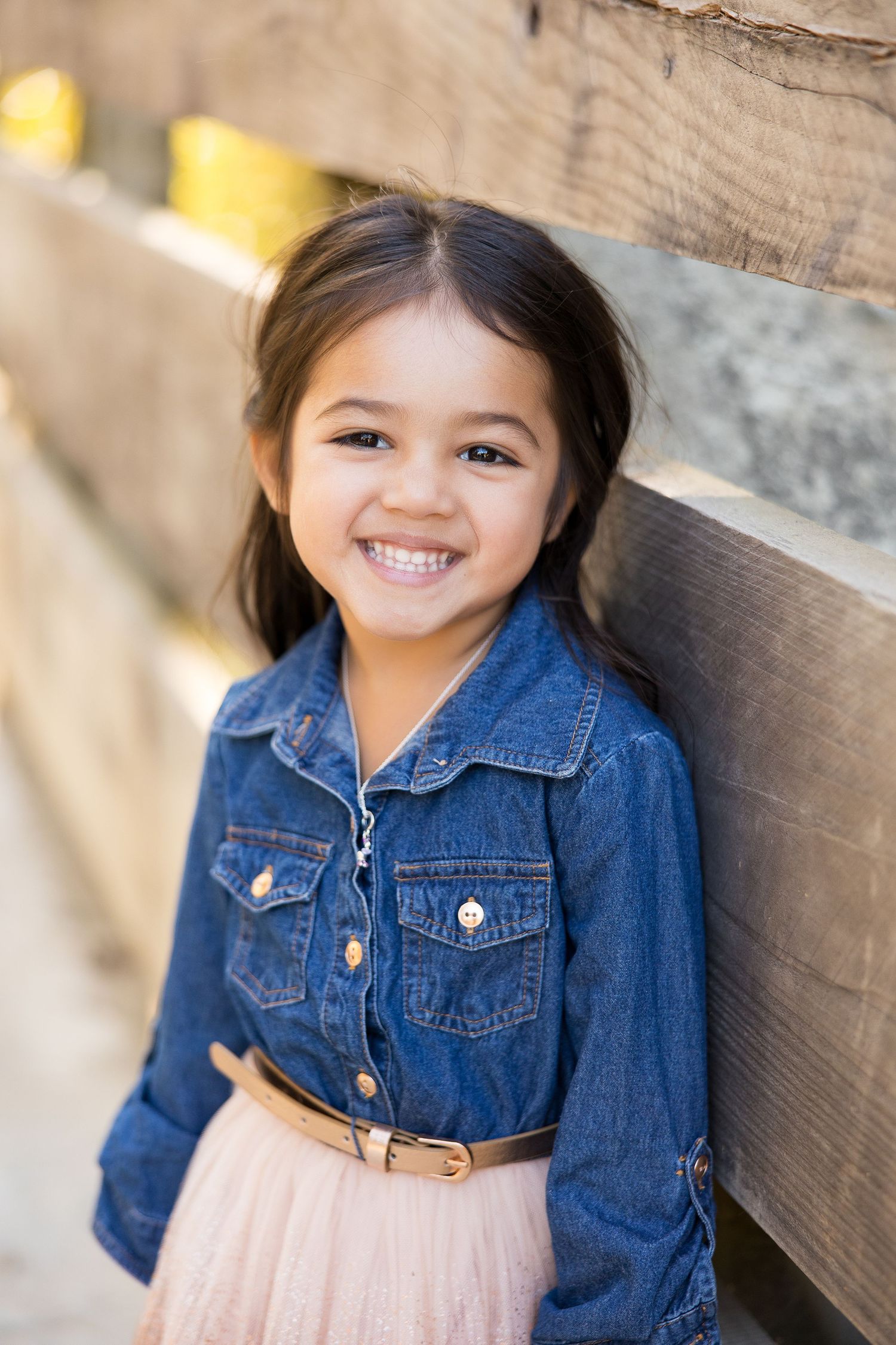 Child in denim jacket smiling against wooden fence background.