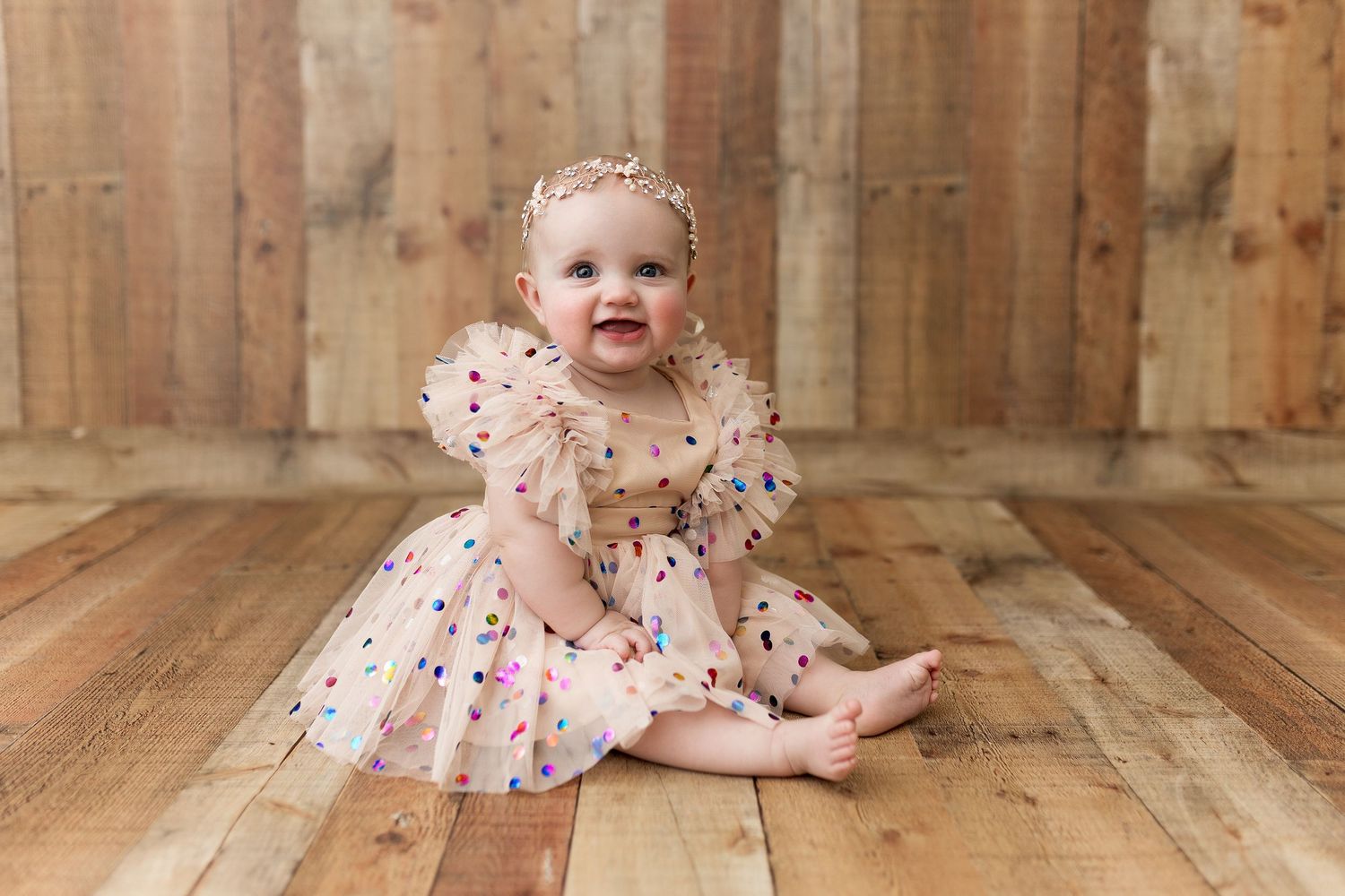 Baby wearing pink tulle dress with polka dots sitting on wooden floor.