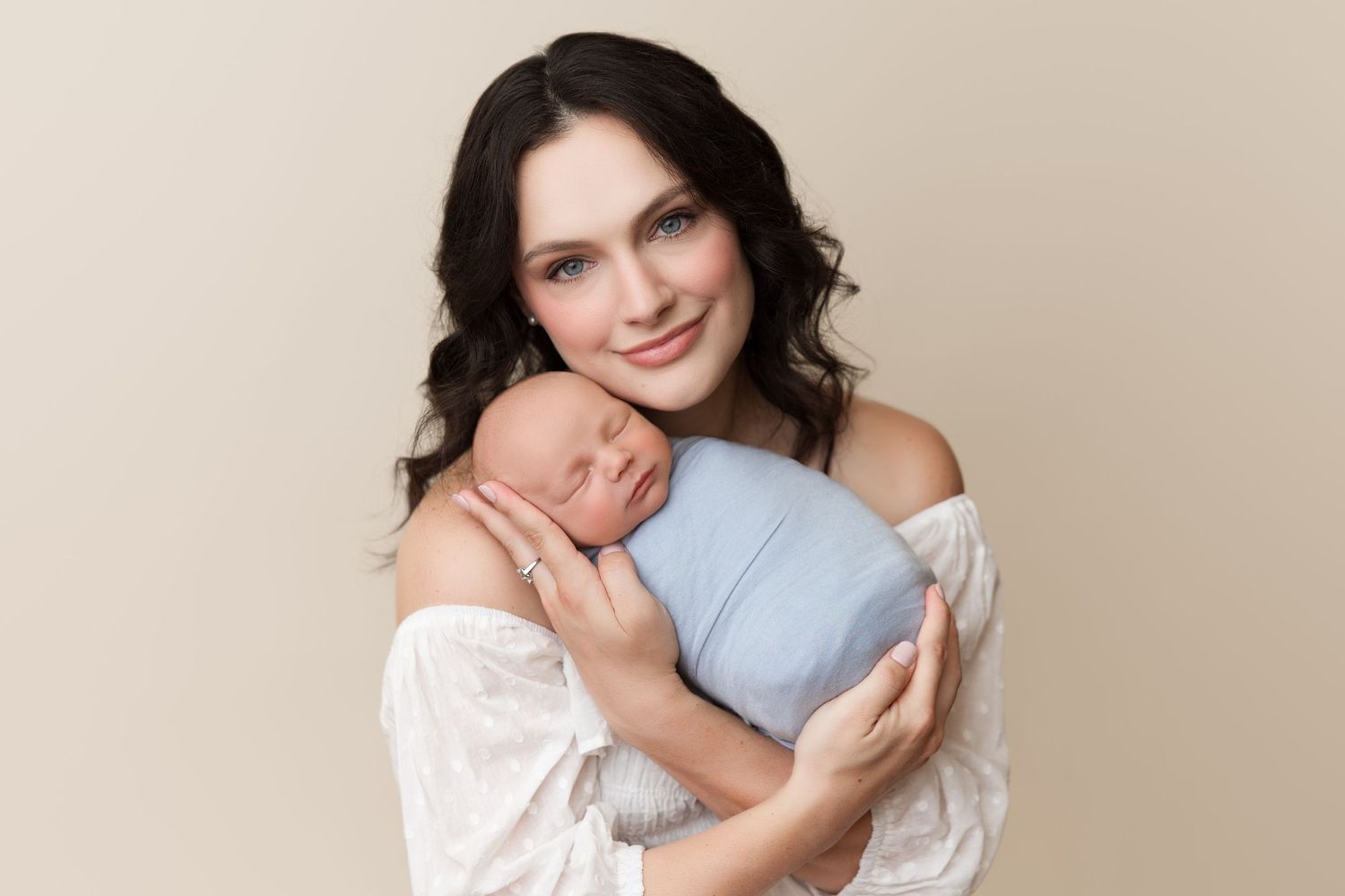 Mother embracing baby wearing blue outfit against beige studio backdrop.
