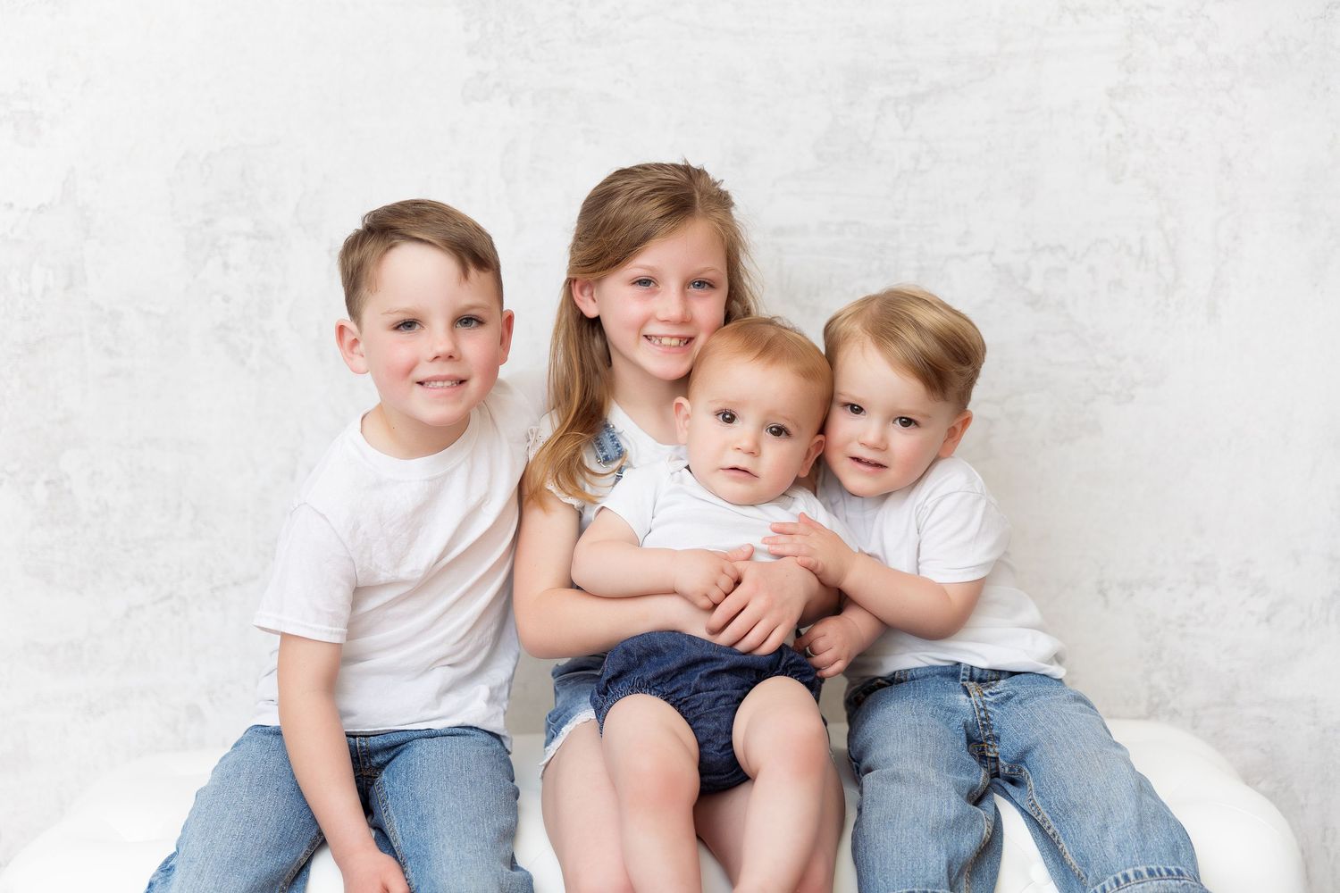 Group of children in coordinated outfits sitting together on white bench for family portrait.