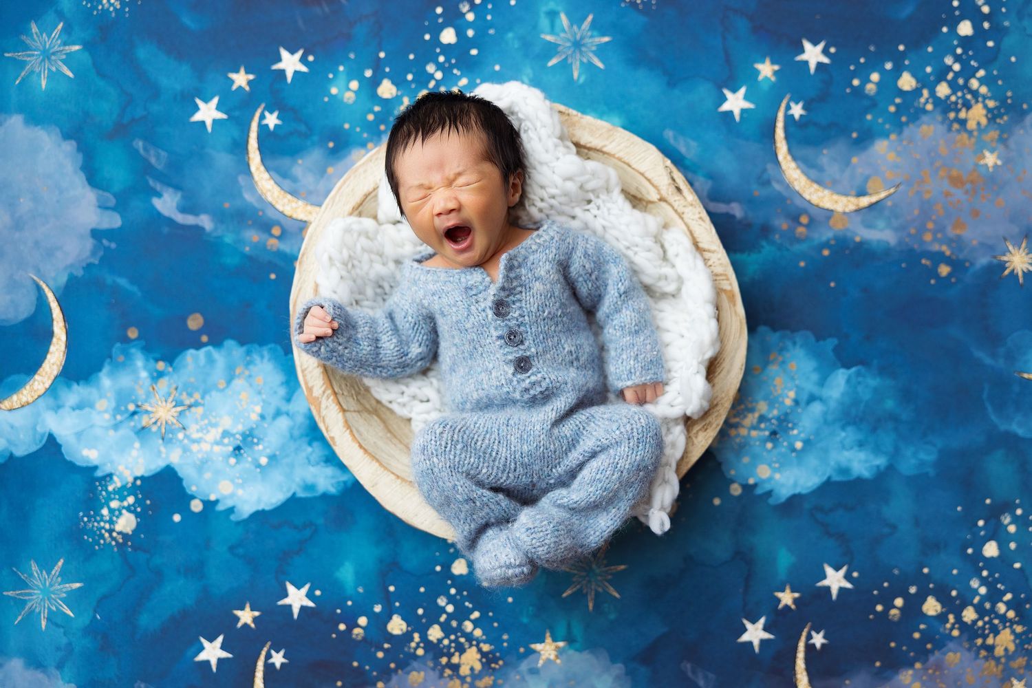 Baby in blue sleeper lying in round basket against starry night backdrop.