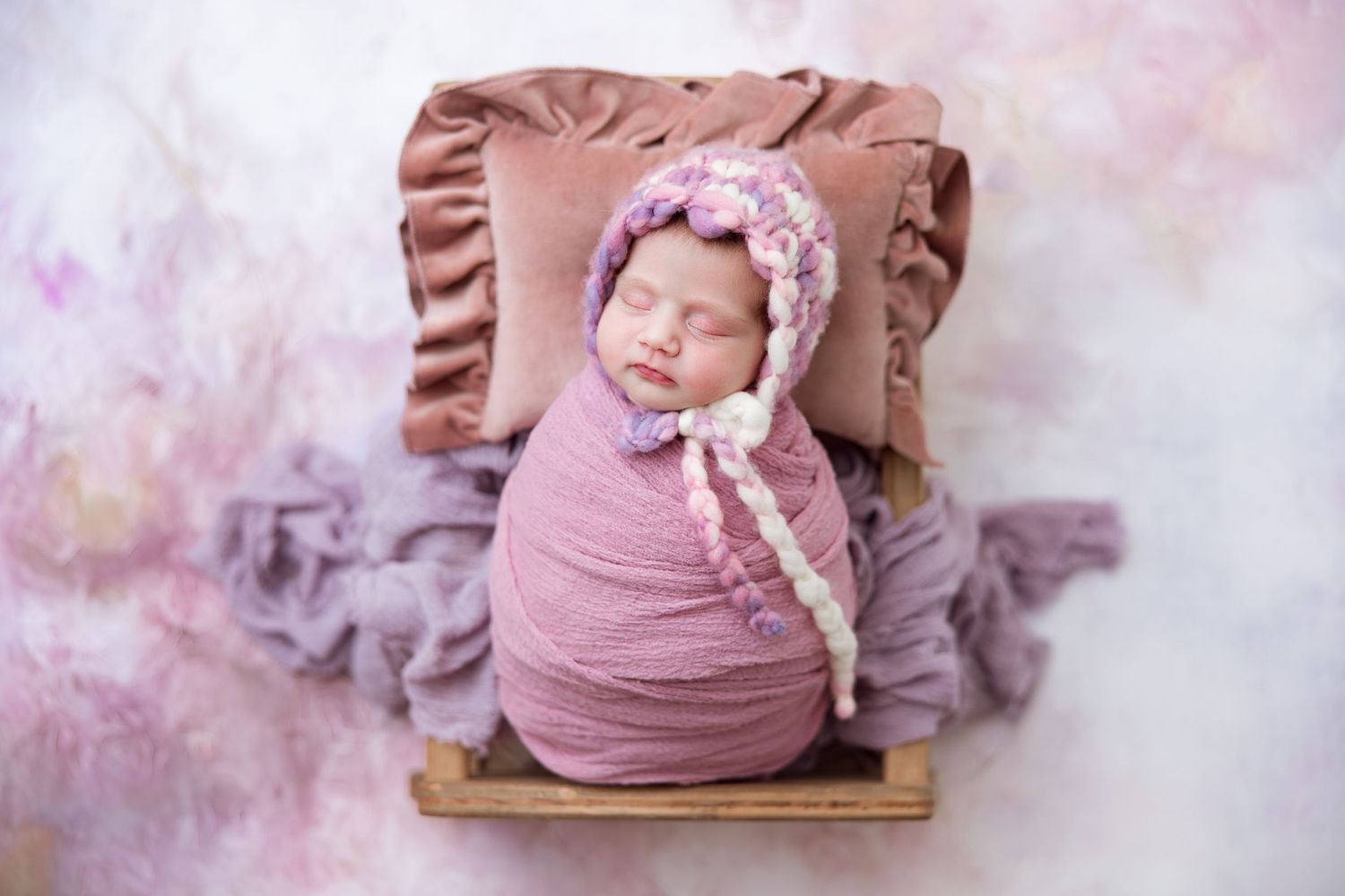Newborn wrapped in mauve swaddle sleeping on ruffled pillow with bonnet.