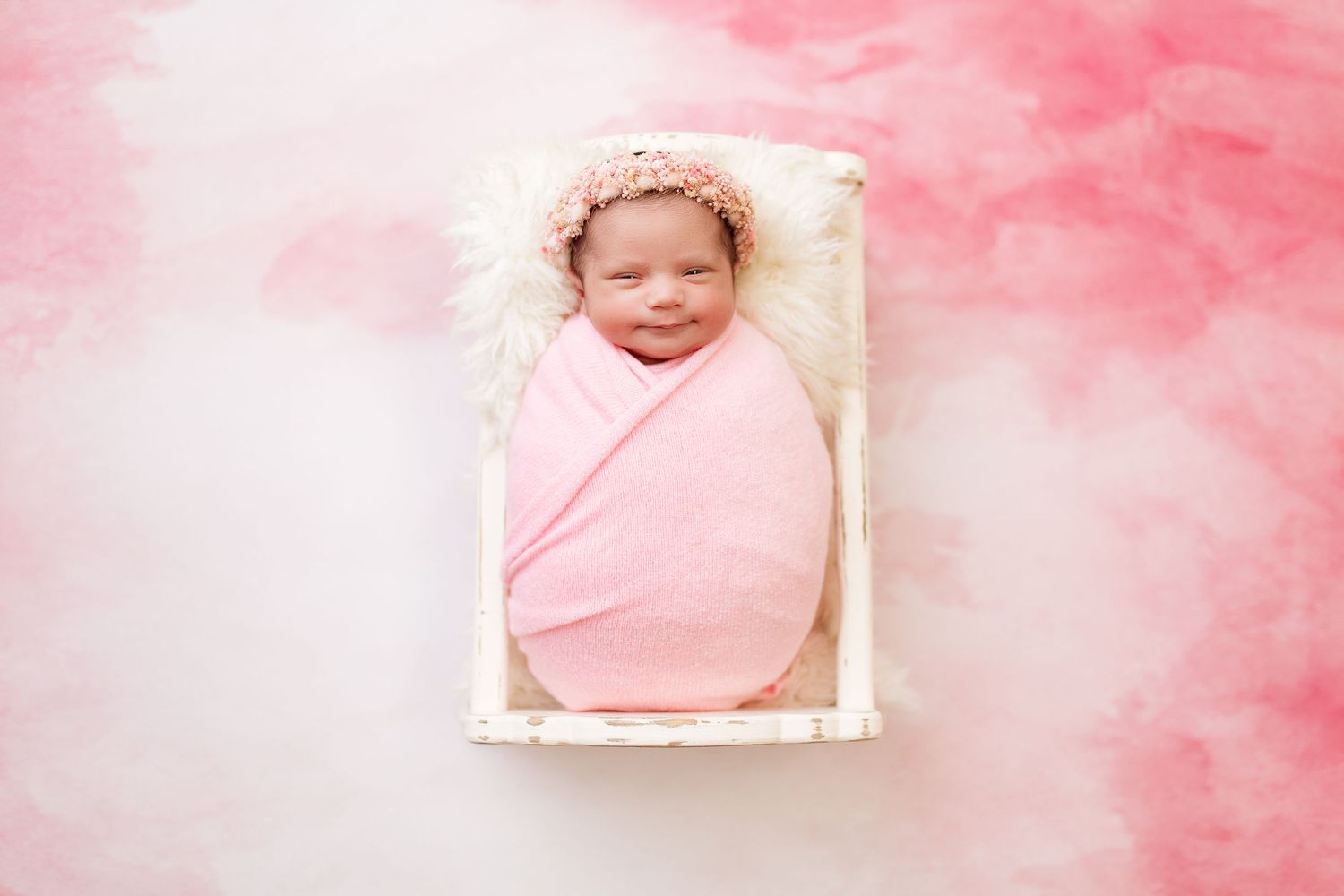 Swaddled baby in pink wrap resting in white basket against pink background.