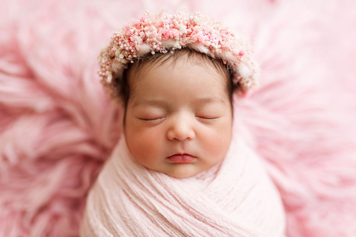 Close-up of sleeping newborn with pink floral crown against soft pink backdrop.