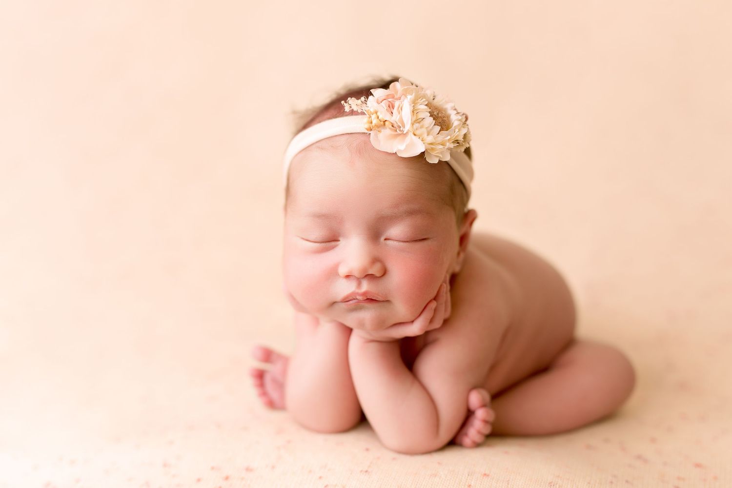 Sleeping newborn posed with head resting on hands wearing peach floral headband.