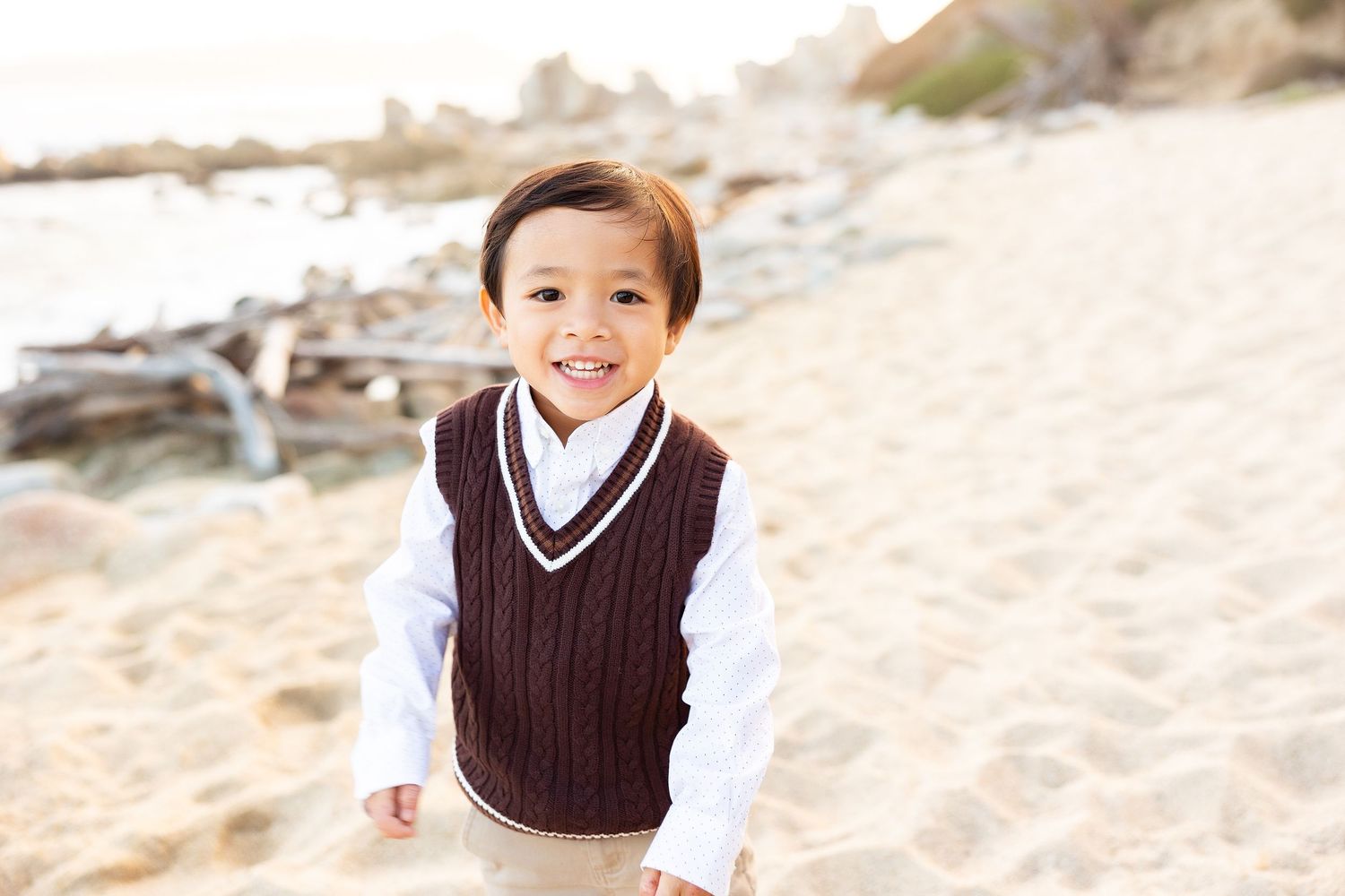 Child in brown sweater vest and white shirt poses happily on beach.