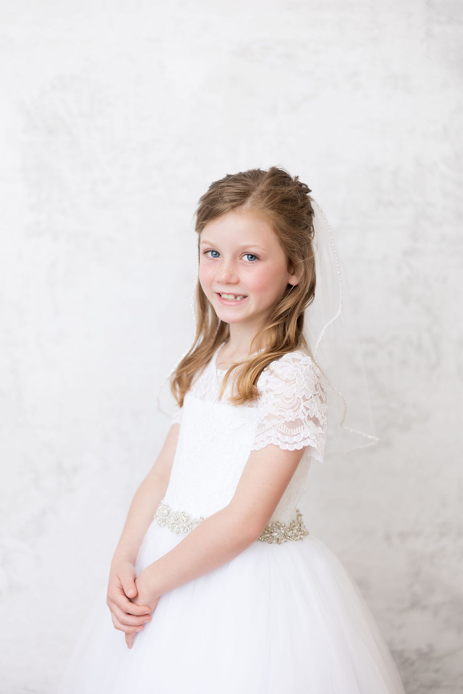 Young girl in white lace First Communion dress smiling against light background.