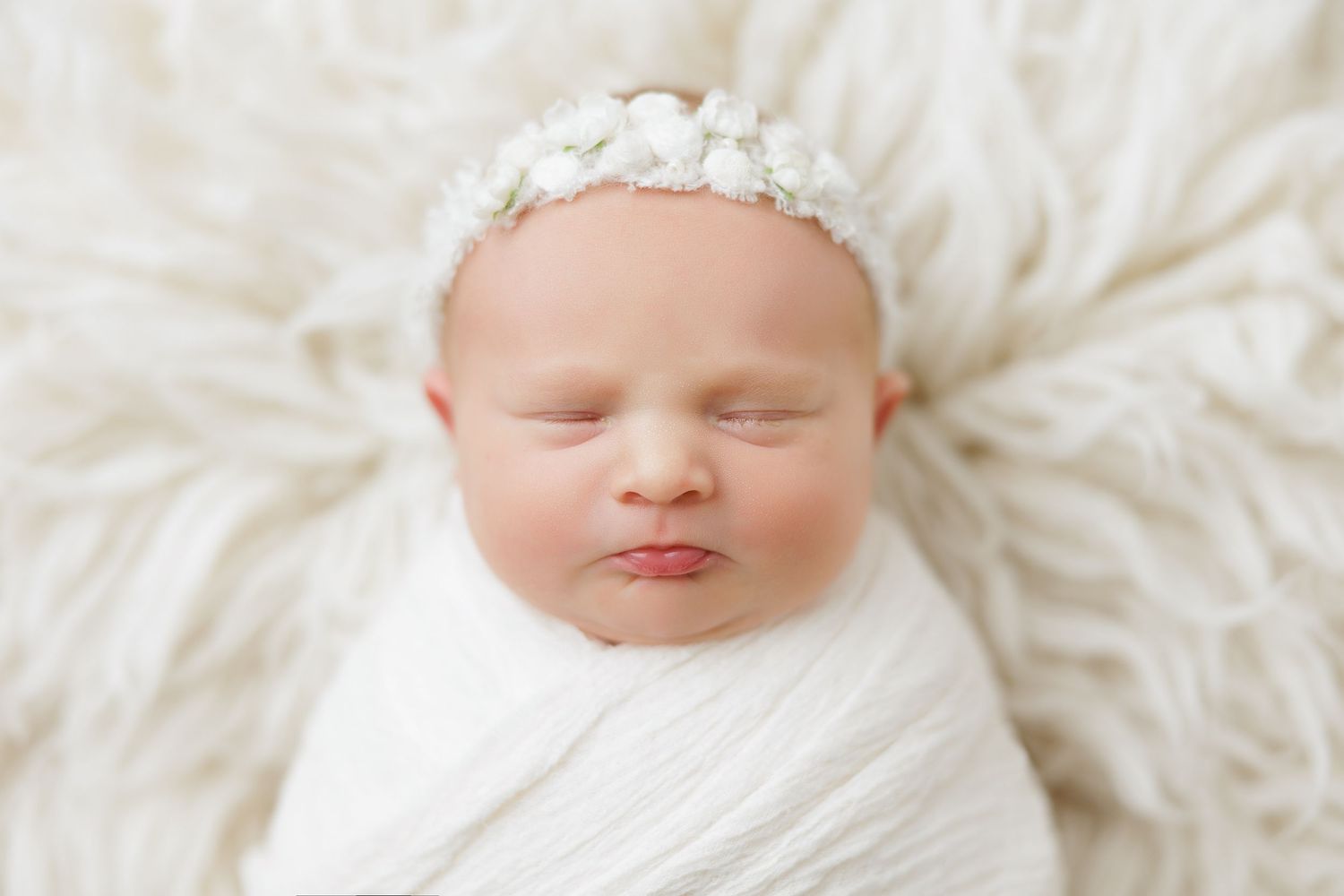Sleeping newborn baby girl wrapped in white swaddle with floral crown on fluffy white fur backdrop.
