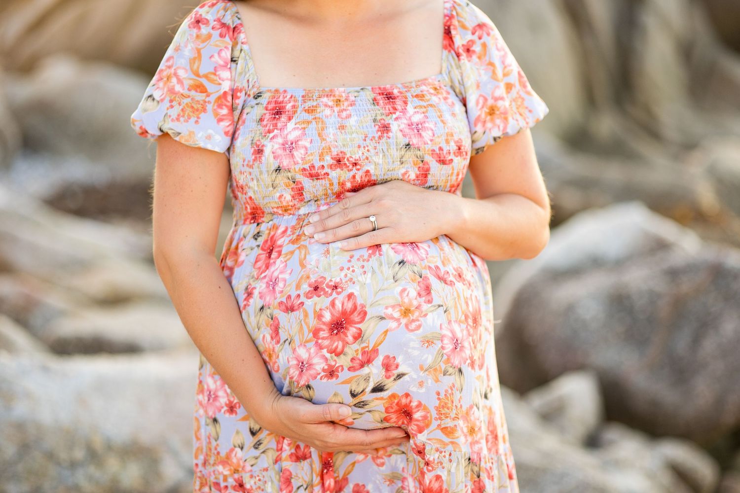 Pregnant woman in floral dress stands on rocky beach during golden hour.