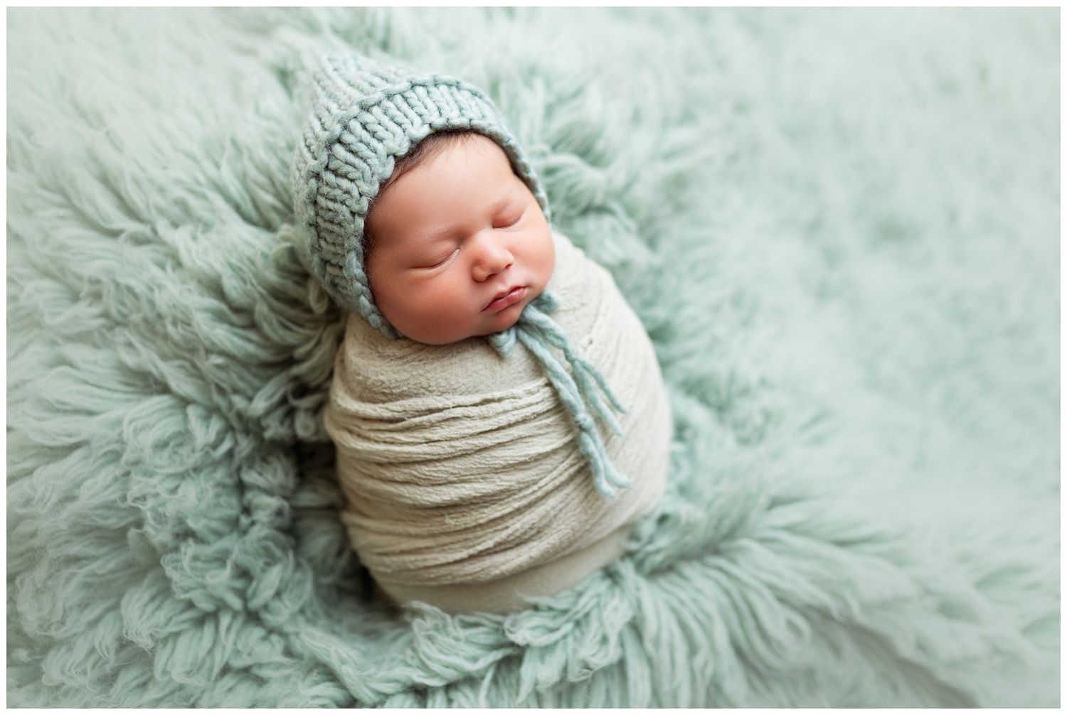 Newborn baby wrapped in soft white fabric sleeping peacefully on mint green textured blanket.
