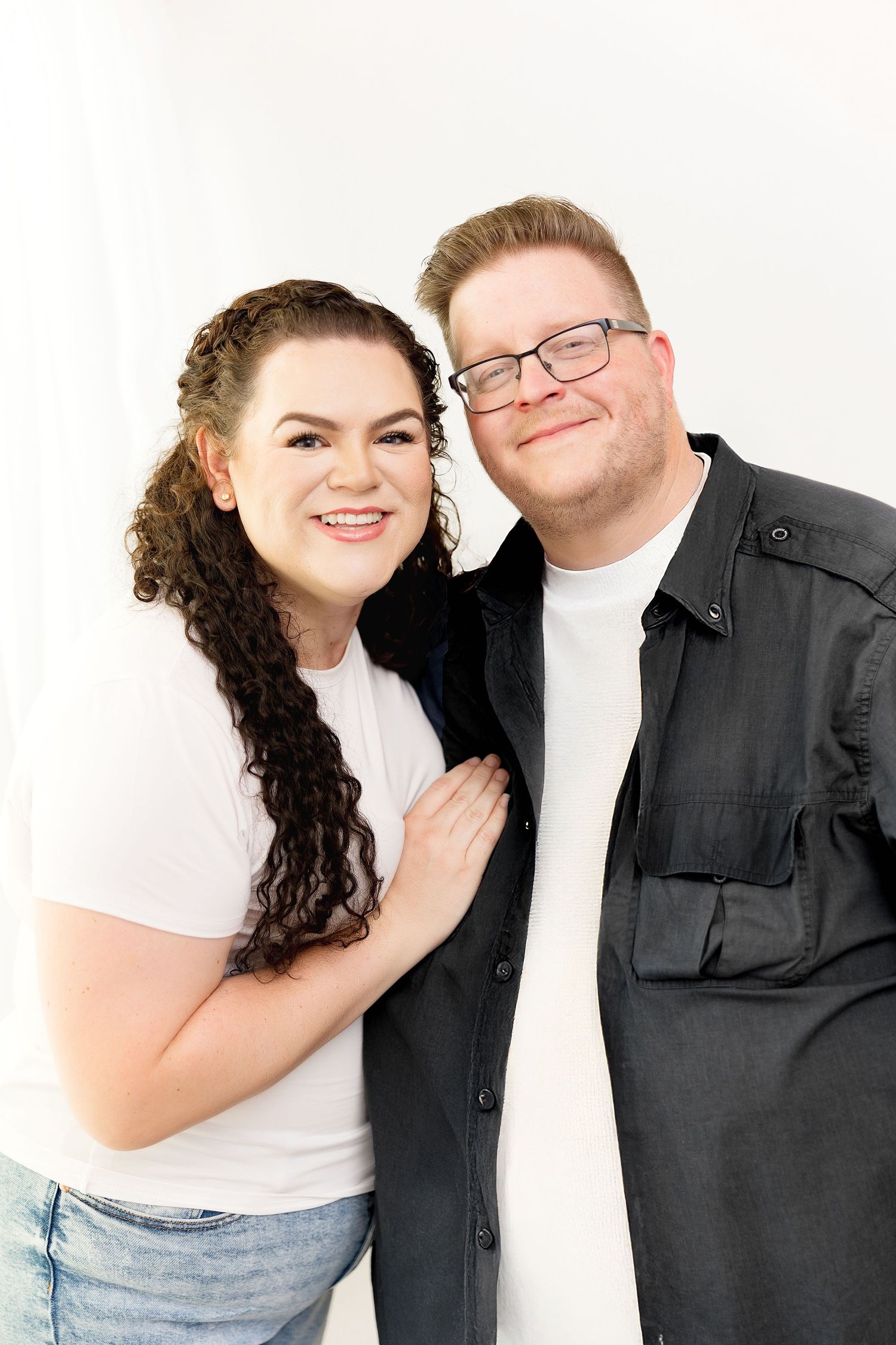 A smiling couple poses together against a white background, woman in white top and man in black shirt with glasses.