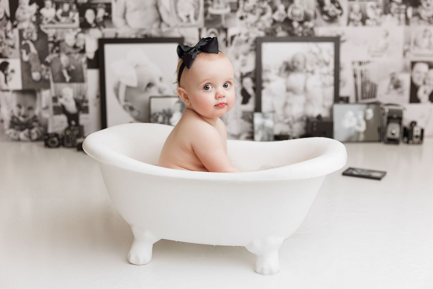 Baby with black bow sitting in decorative white bathtub against vintage photo wall.