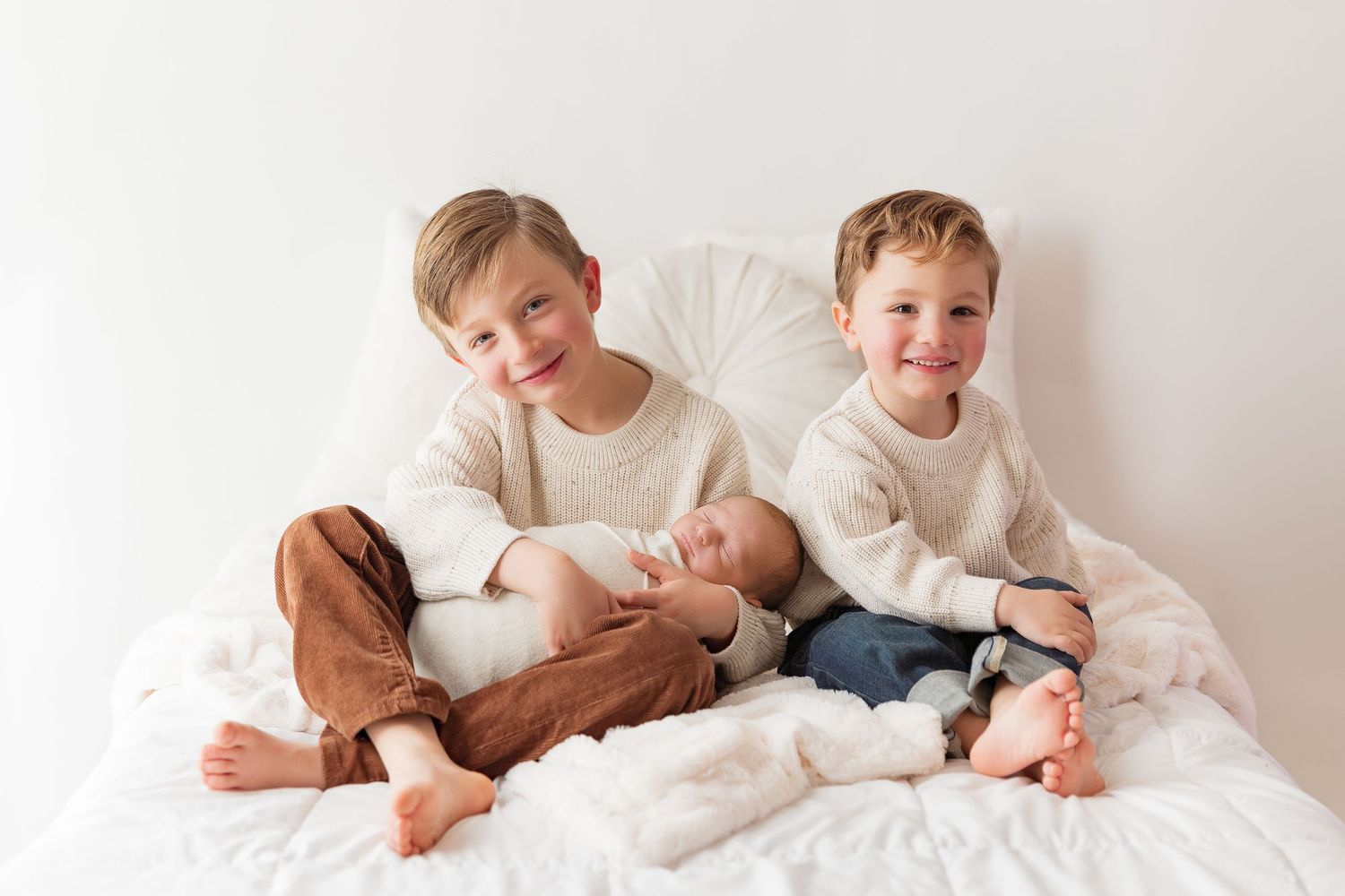 Two children in white sweaters sit with sleeping baby on white bed.