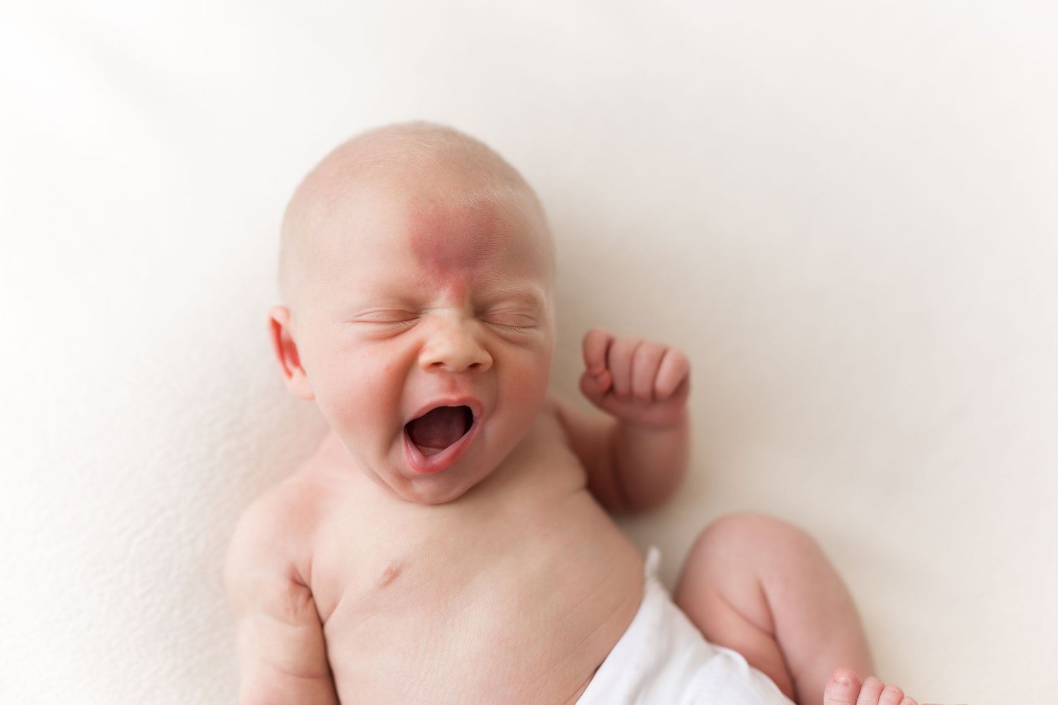 Baby in white clothing yawning or crying against white background.