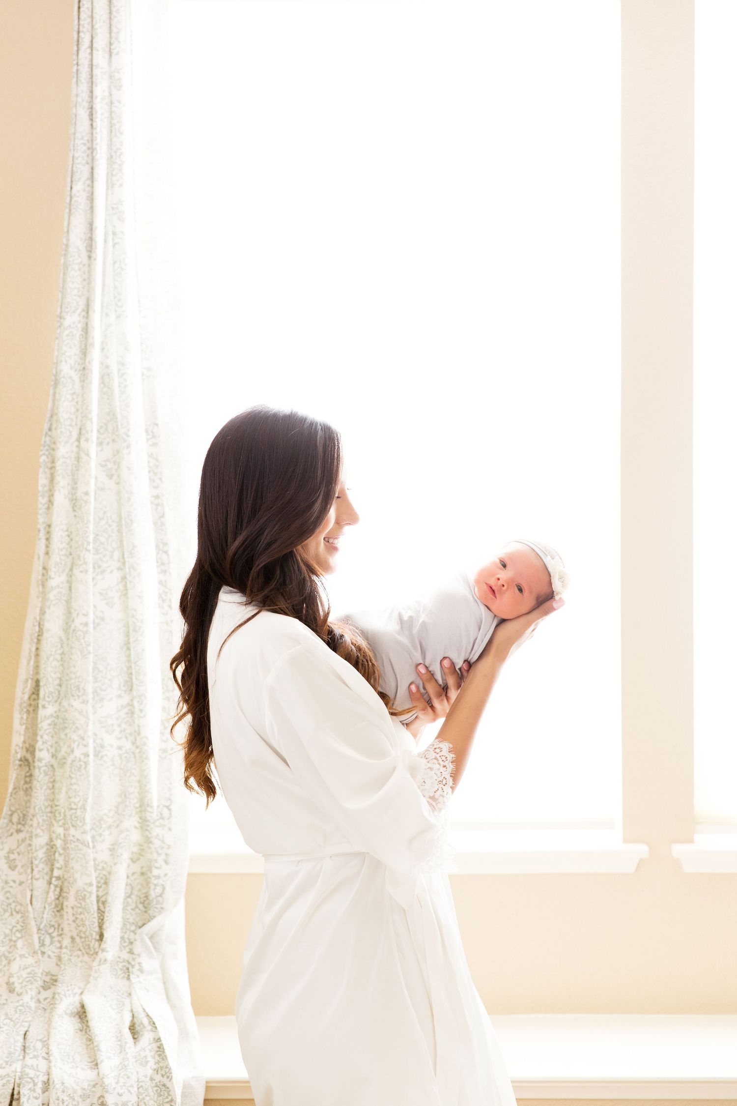 Mother in white robe holds newborn baby by sunlit window with sheer curtains.