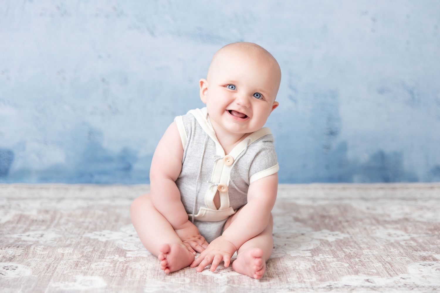 Happy baby in light-colored romper sitting on wooden floor with blue backdrop.