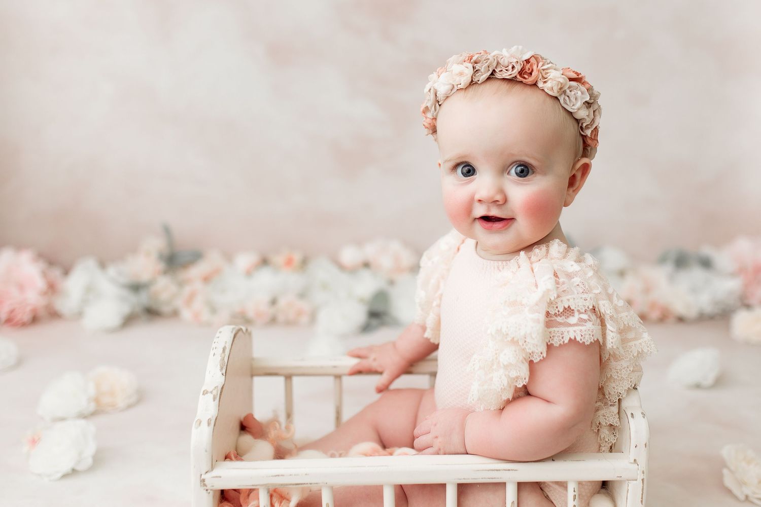 Baby in lace romper sitting in white vintage crib with pink floral decorations.
