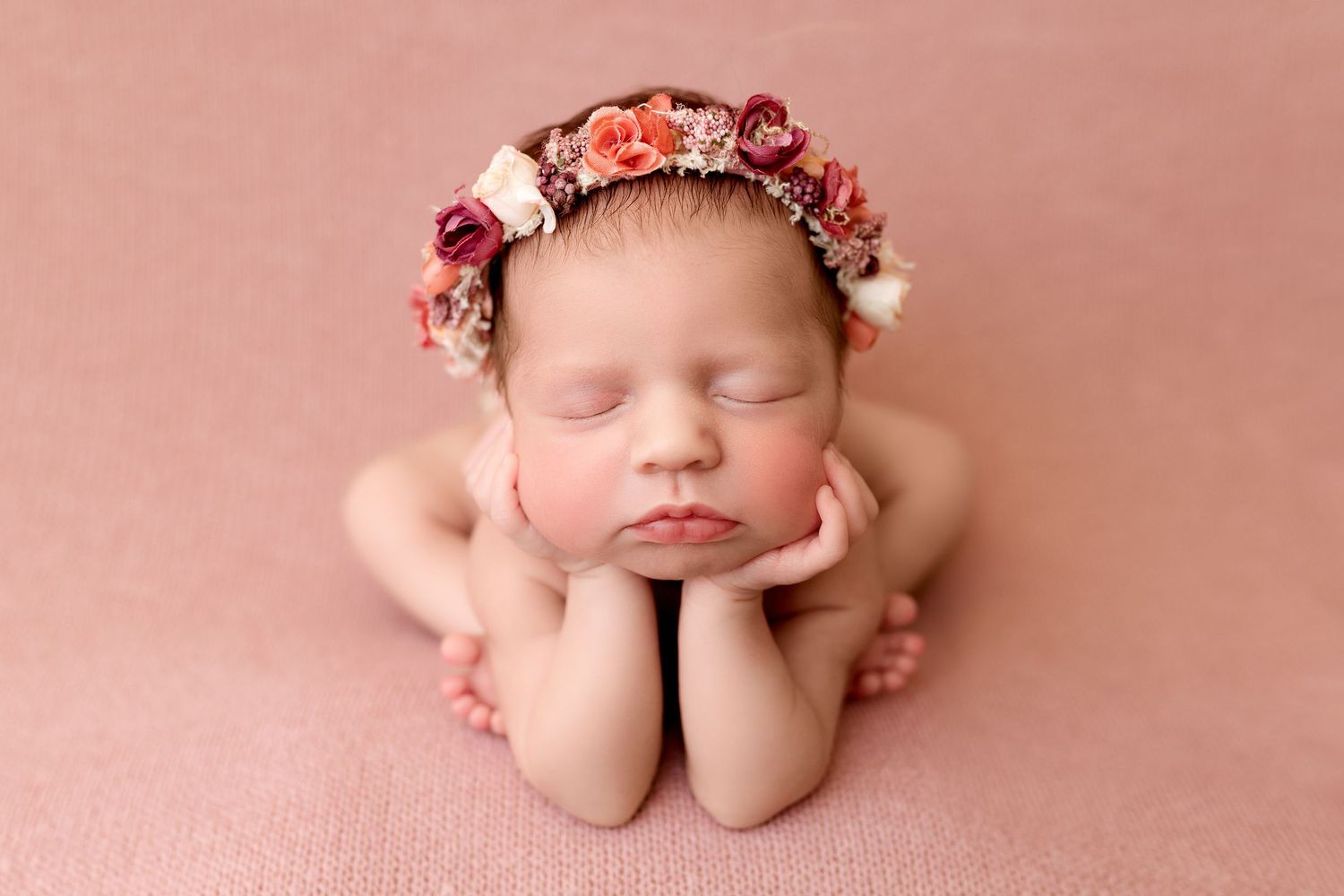 Sleeping newborn wearing floral crown posed in froggy position on pink backdrop.