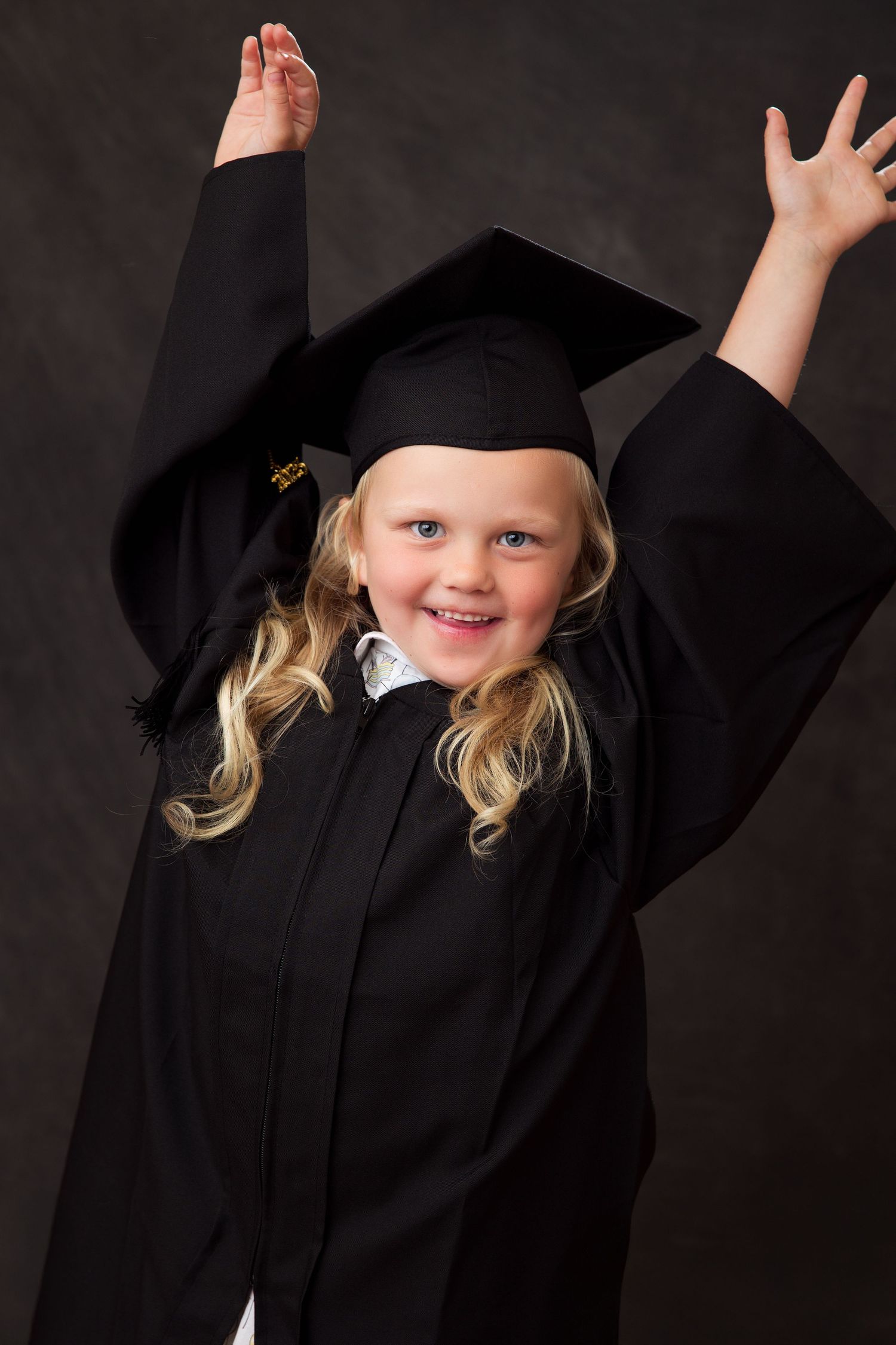 A joyful young graduate in black cap and gown raises their arms in celebration against a dark background.