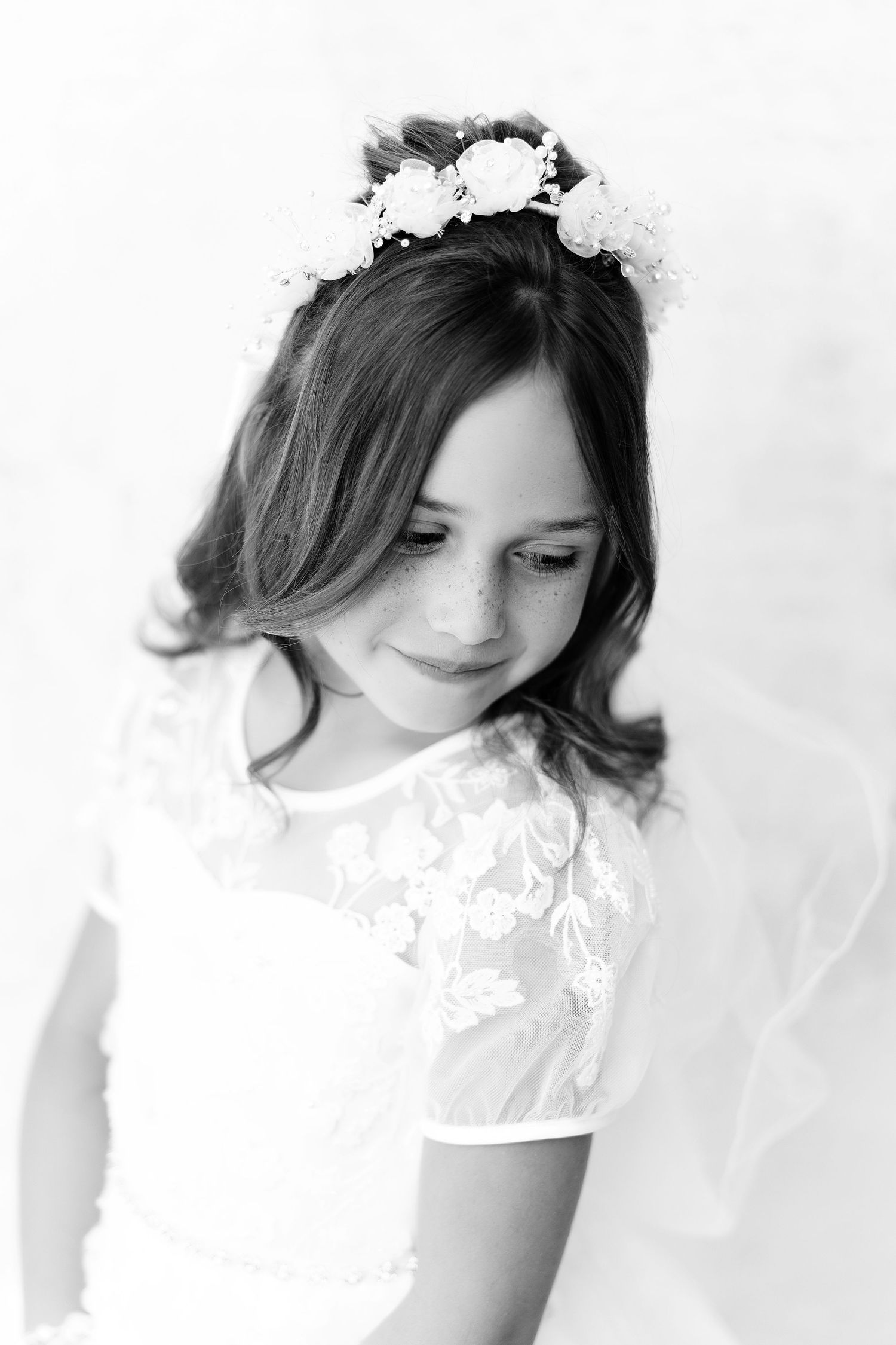 Black and white portrait of someone wearing a white lace dress and floral crown looking down with a gentle smile.