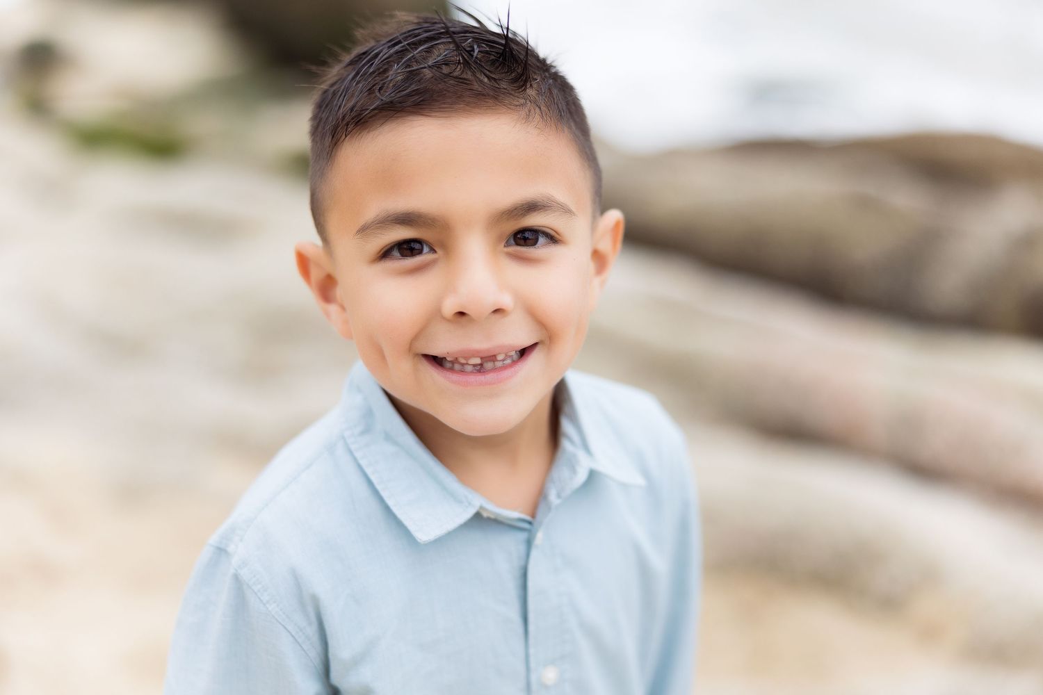 Young boy in light blue polo shirt smiles at beach with rocks in background.