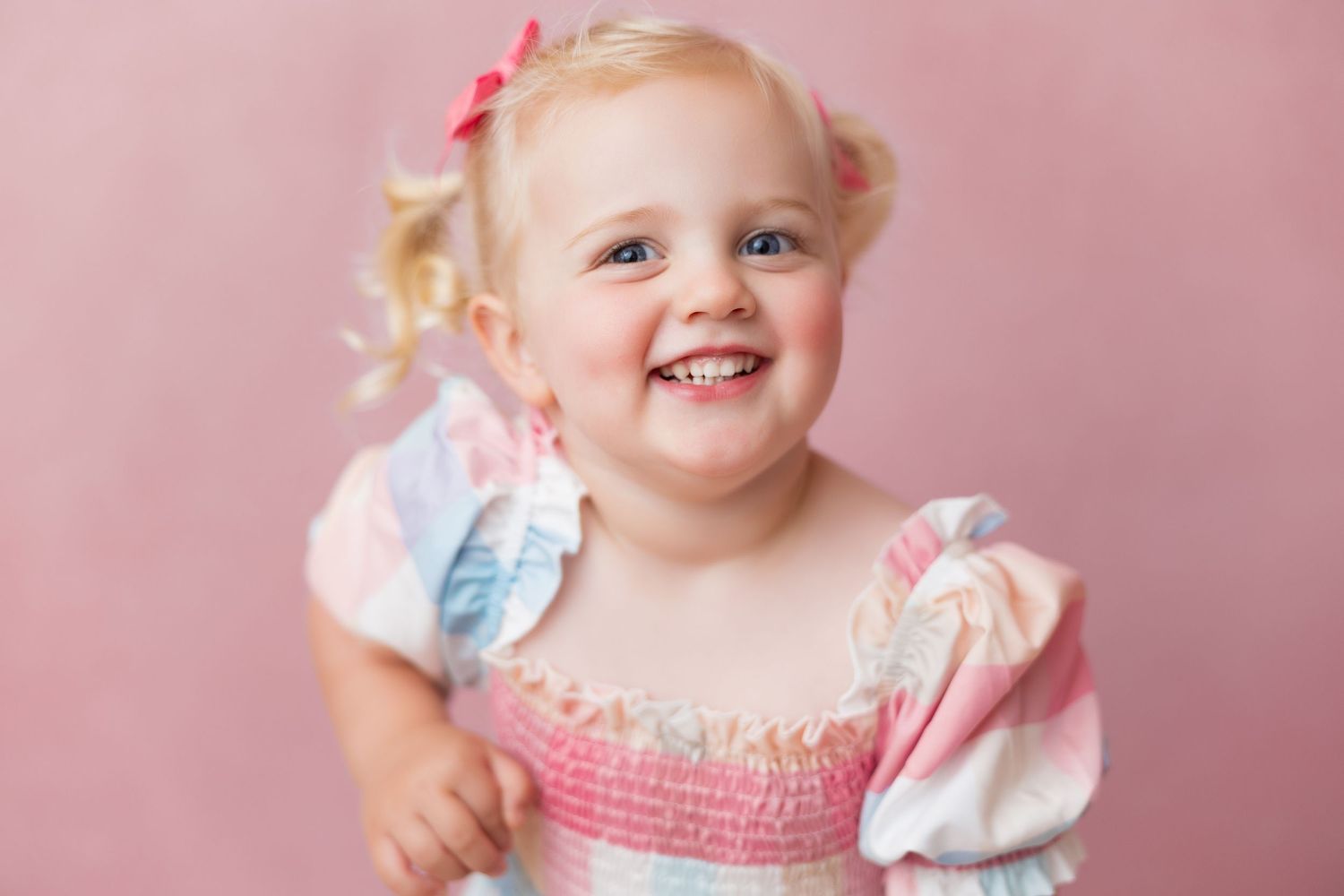 A happy young toddler in a pink and white dress smiles brightly against a rosy pink backdrop.