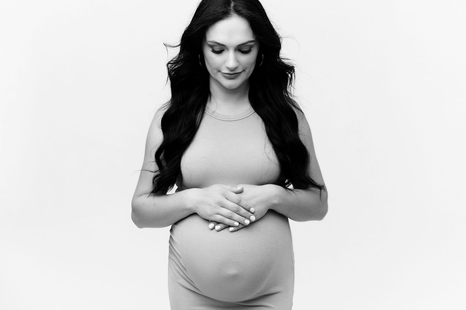 A black and white portrait of an expectant mother in a fitted dress holding her baby bump with downcast eyes.