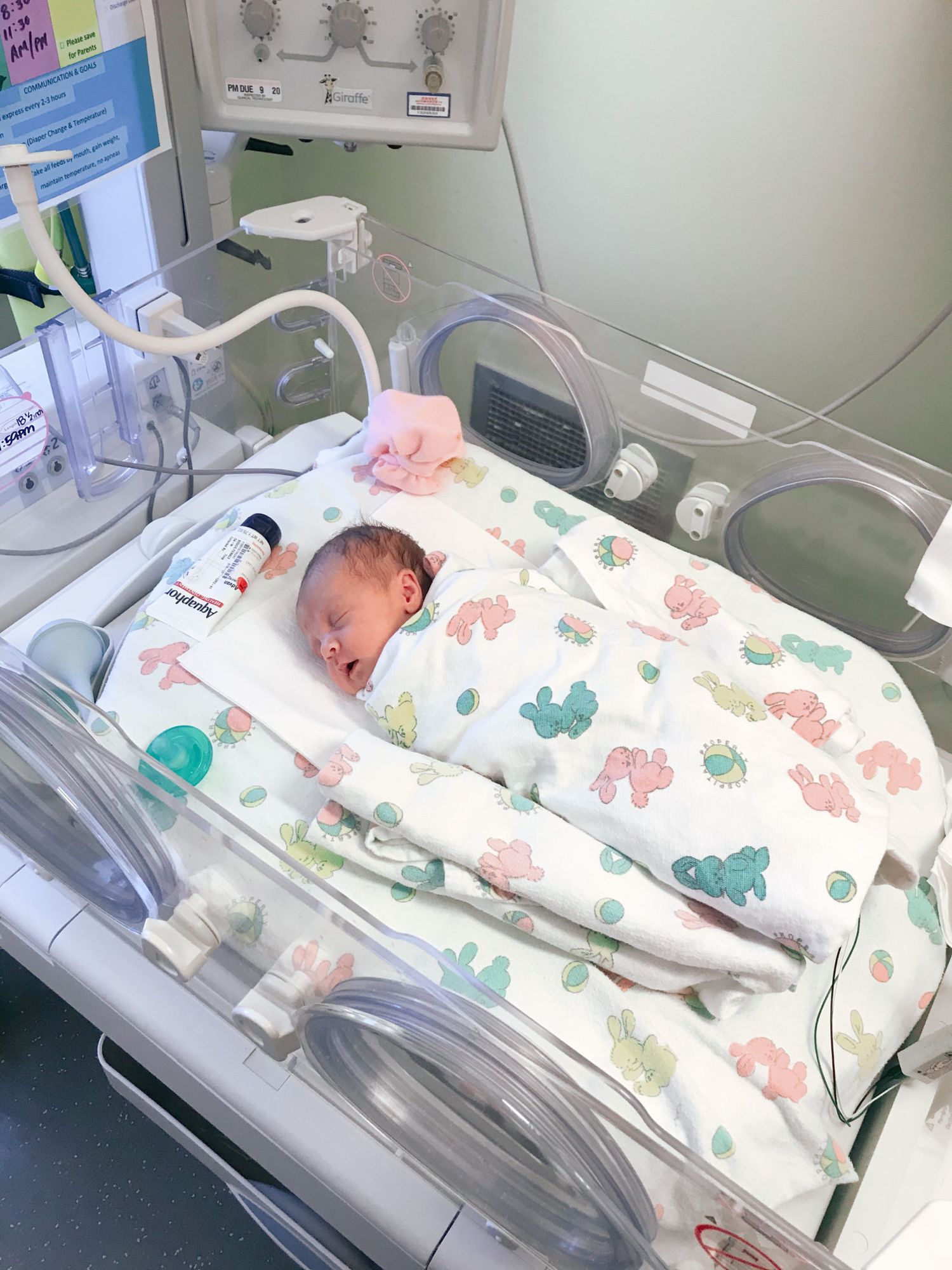 A newborn baby sleeping in a hospital bassinet covered with a blanket featuring colorful dinosaur patterns.