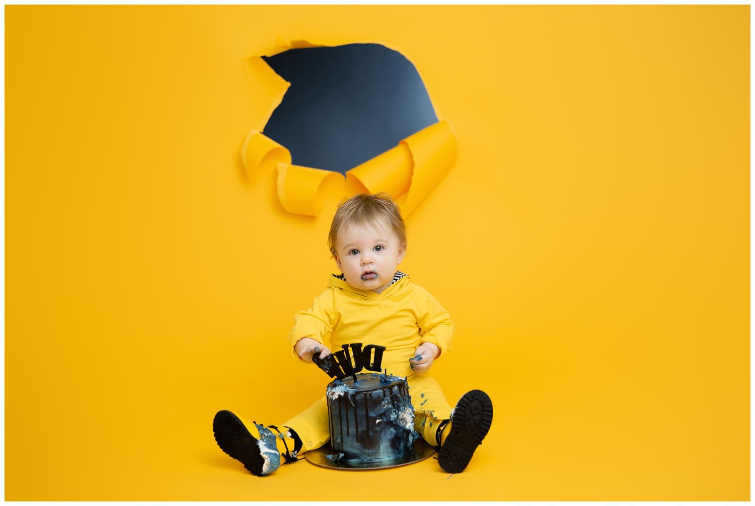 Baby wearing yellow shirt and graduation cap sits on yellow background in creative portrait series.