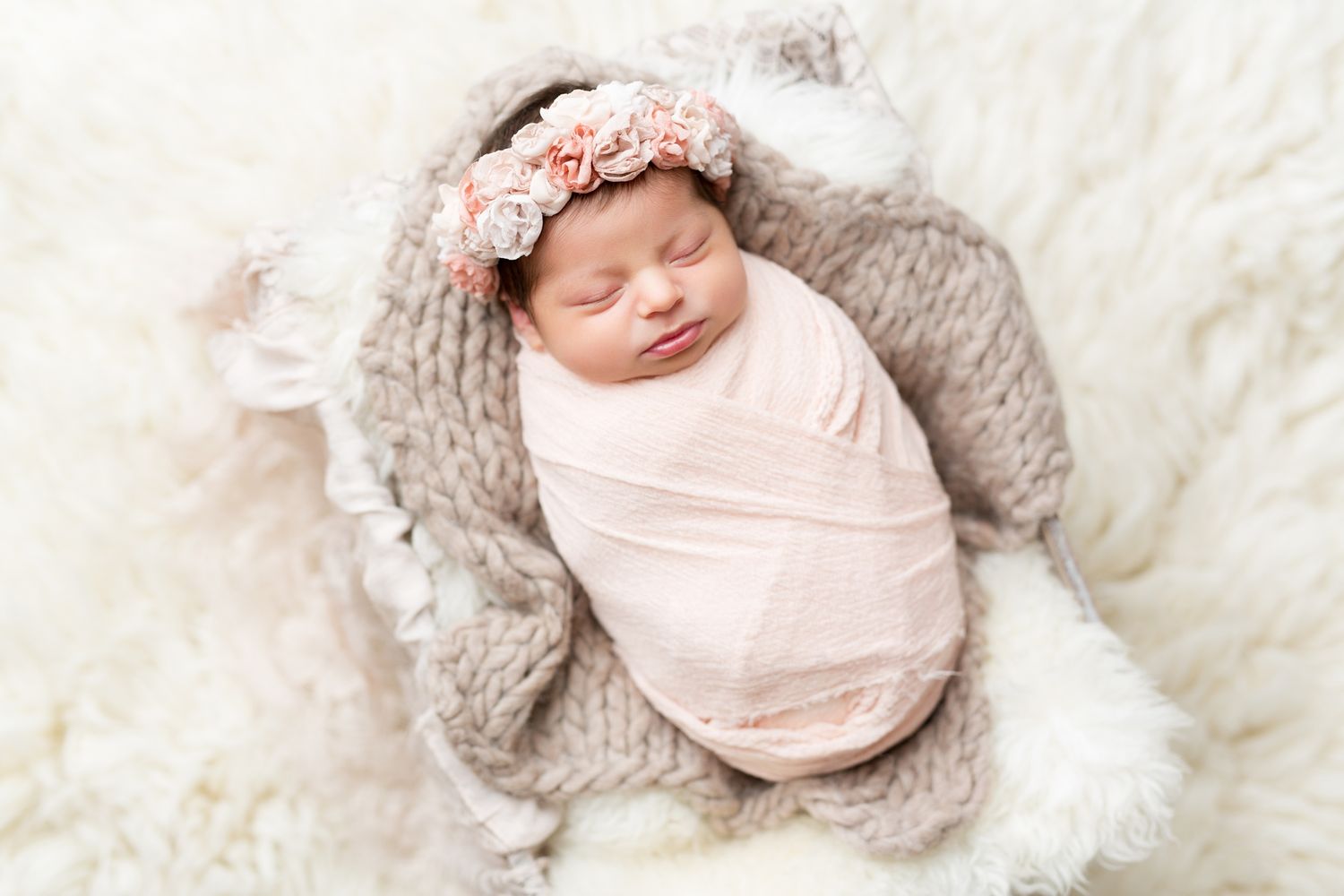 Newborn baby wrapped in pink swaddle blanket sleeping peacefully on soft white fur backdrop.