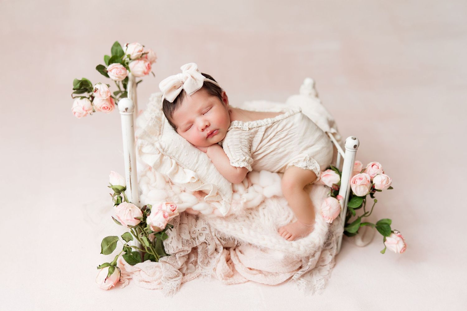 Newborn baby sleeping peacefully among white flowers and soft pink fabric in a professional studio setting.