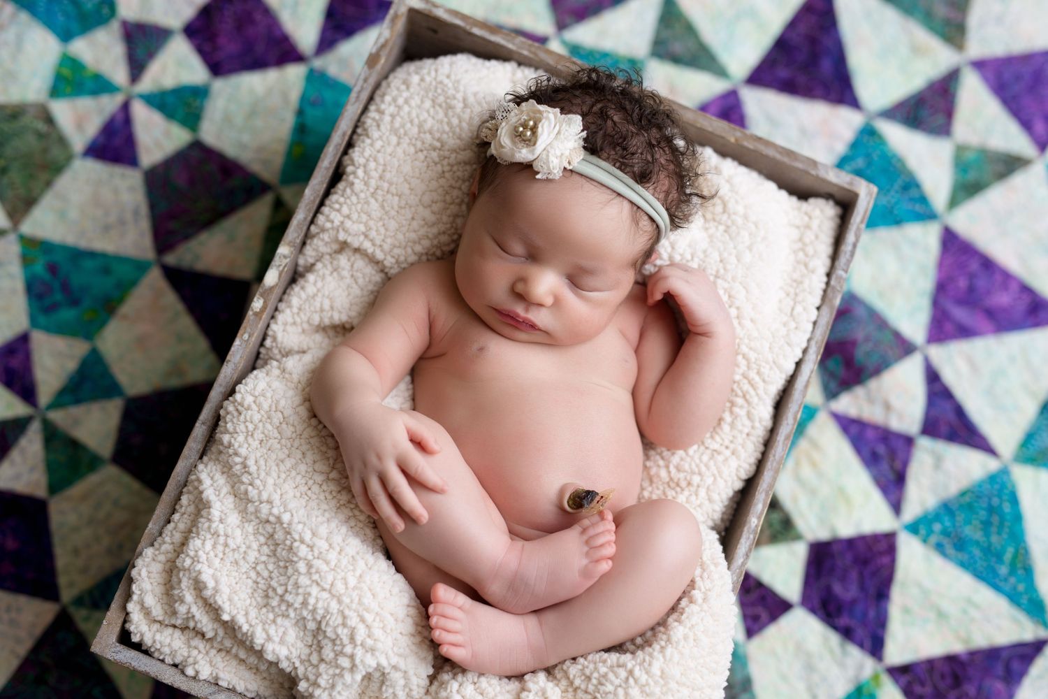 Newborn baby rests on white blanket against colorful geometric patterned background in purple and teal.