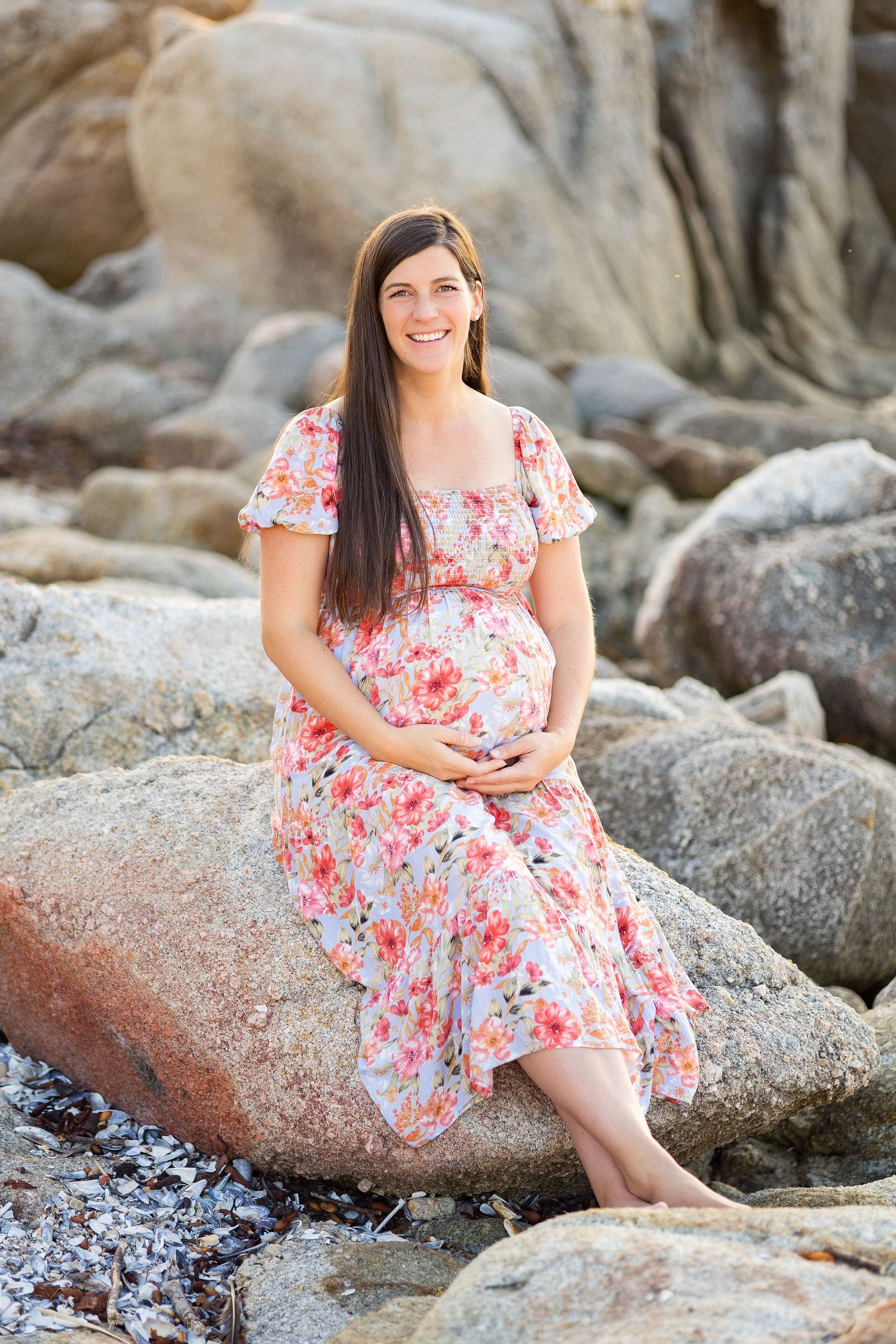 An expectant mother in a floral dress sits on coastal rocks during a sunset photoshoot.