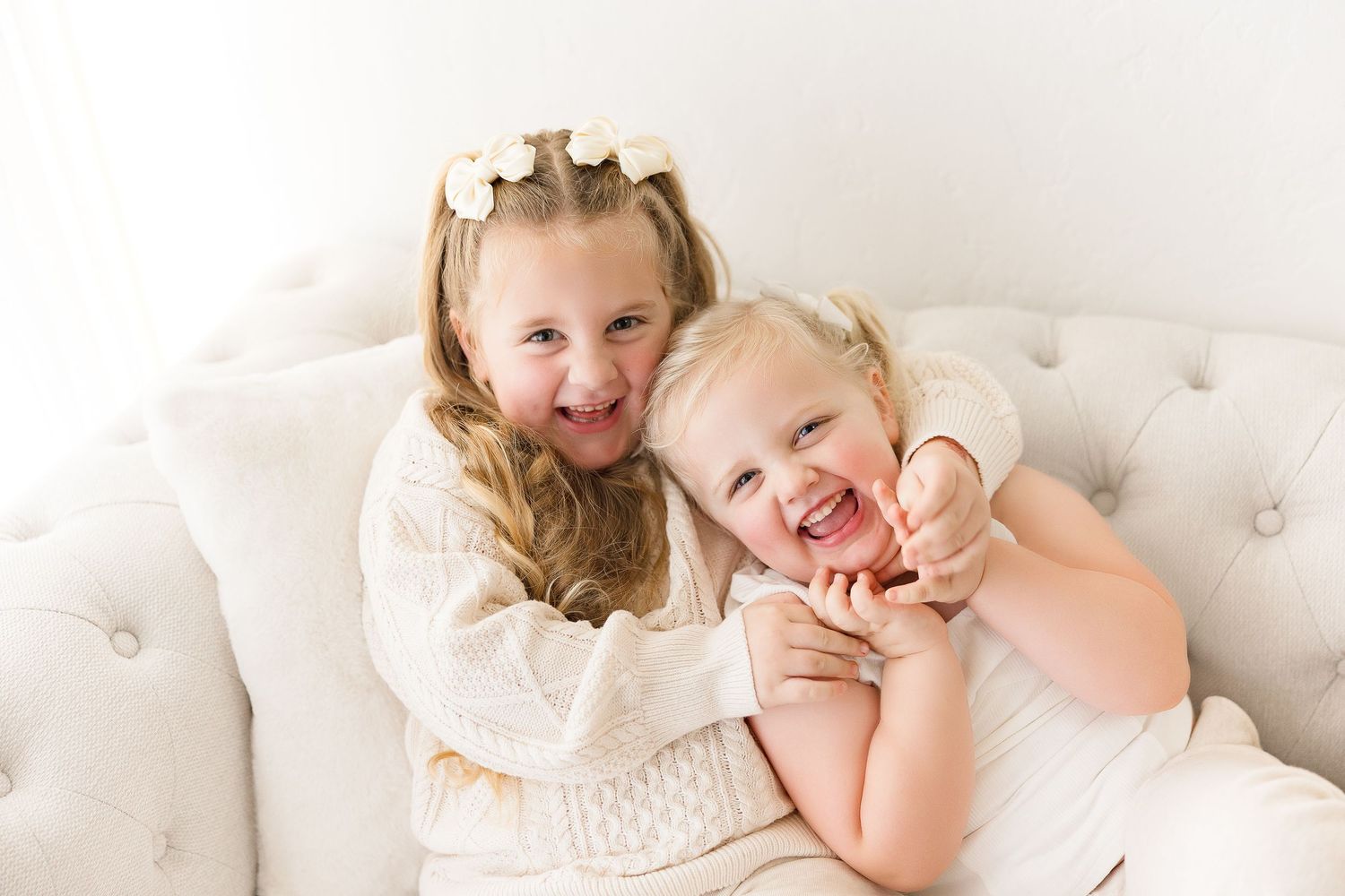 Two children in cream colored outfits laughing and playing together on a white tufted couch.
