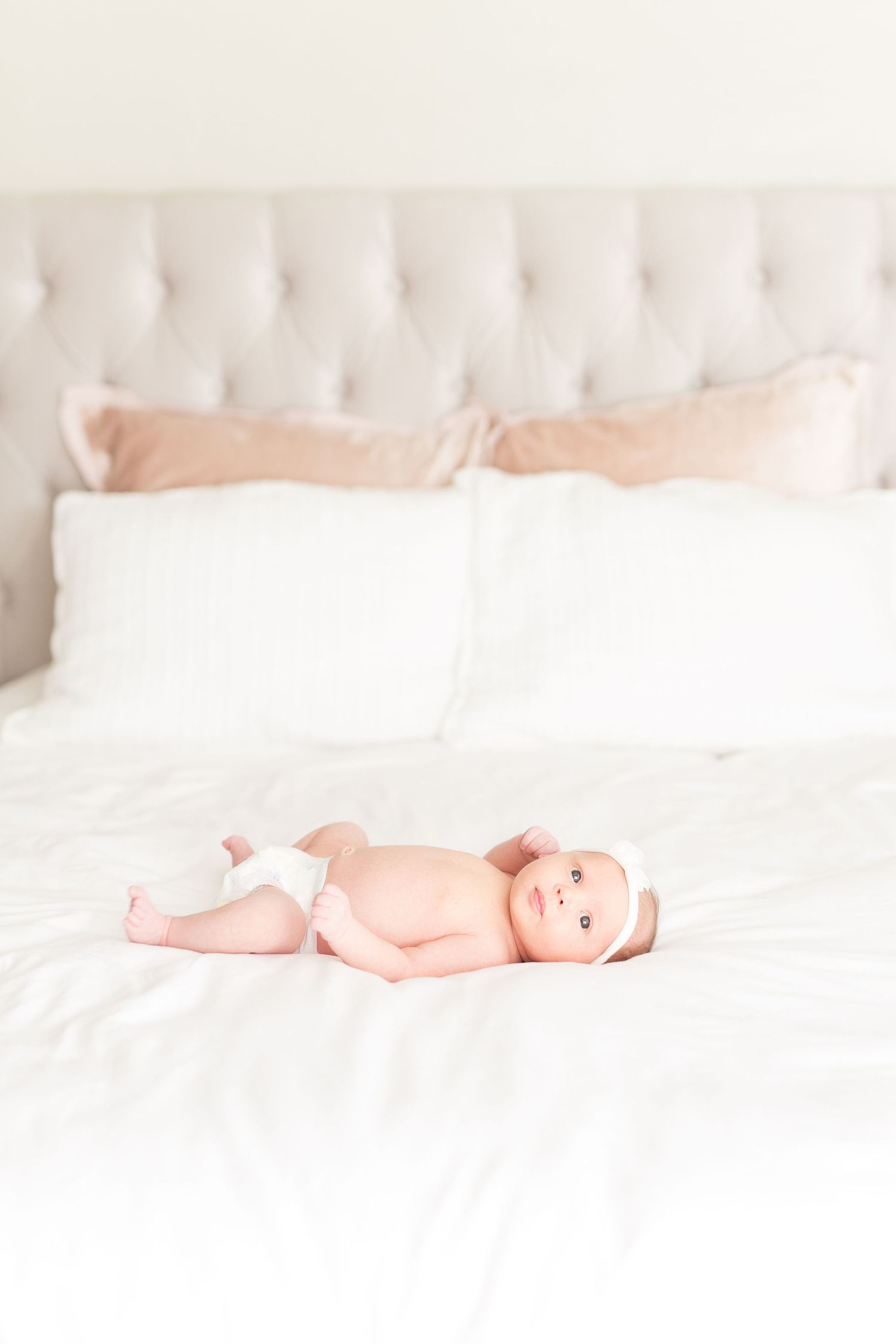 Newborn baby lying on white bedding against tufted headboard background.