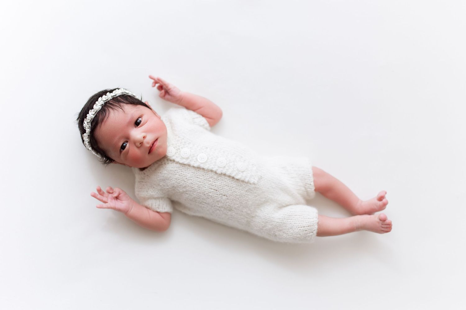 Newborn in white lace outfit lying peacefully against white background.