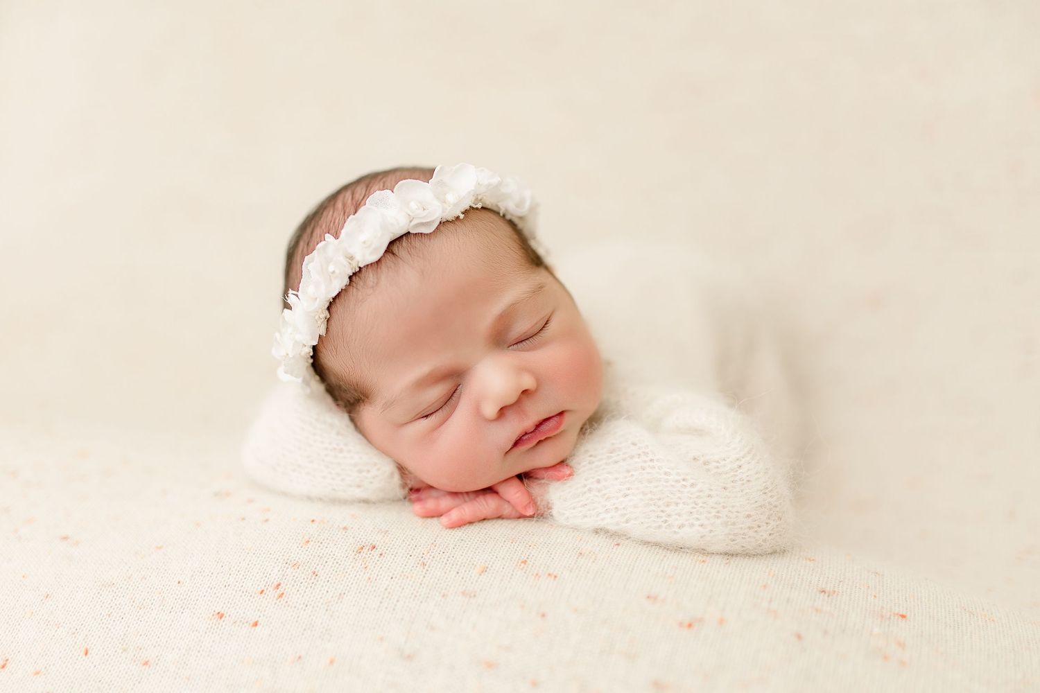 Sleeping newborn wearing lace headband resting on white blanket with peachy tones.